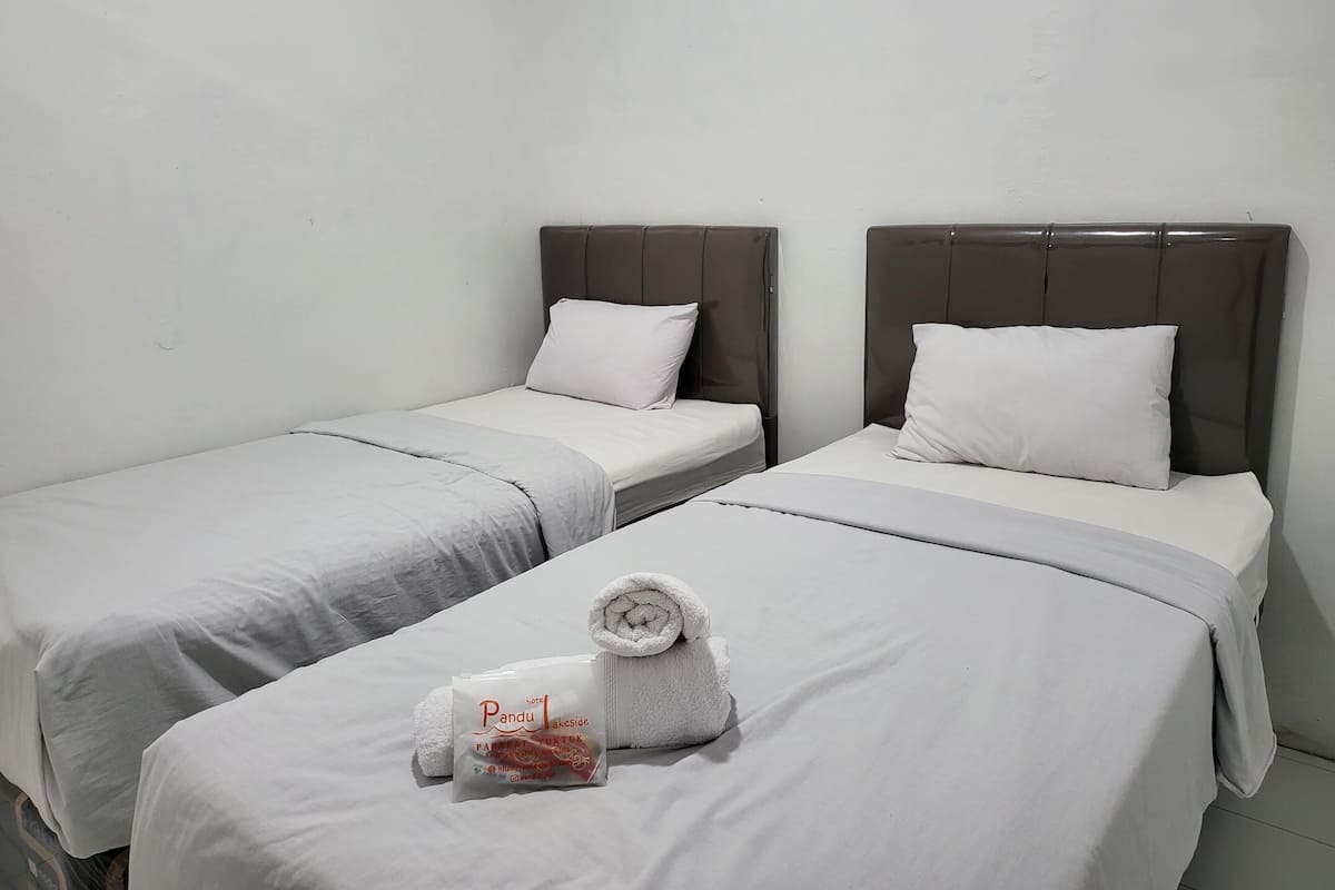 A hotel room with two single beds, each with a grey headboard and white bedding. A towel and a small package are placed on the bed in the foreground.
