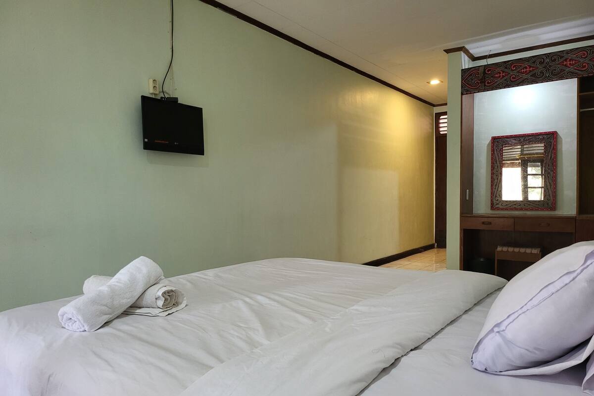 A neatly made bed with white linens and a rolled towel, a flat-screen TV mounted on the wall, and a wooden cabinet with a mirror above it.