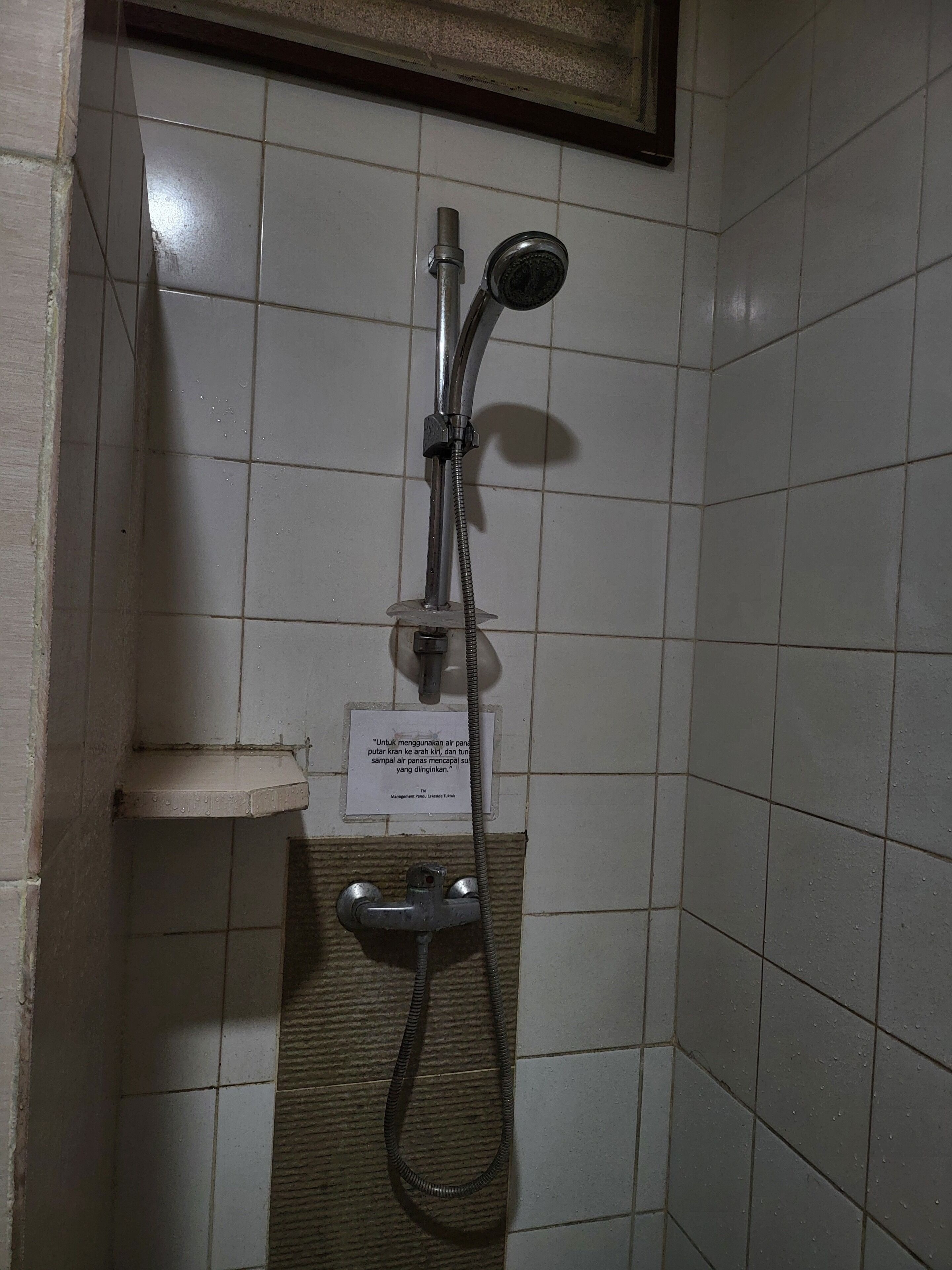A shower with a handheld showerhead and a wall-mounted faucet, a sign with text, and a tiled wall.