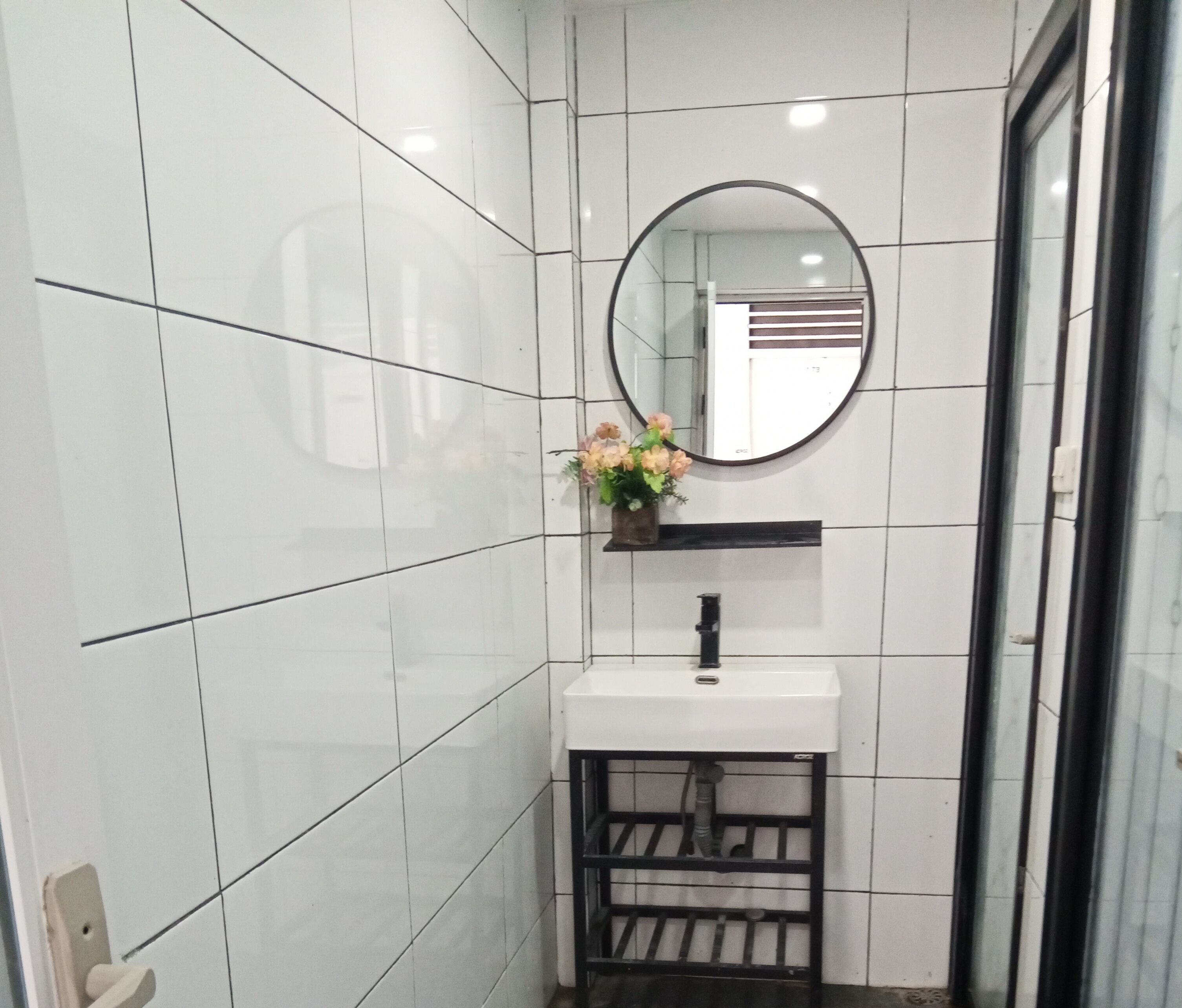 Un baño con paredes de azulejos blancos, un espejo redondo, un lavamanos blanco y una repisa negra con un jarrón de flores.