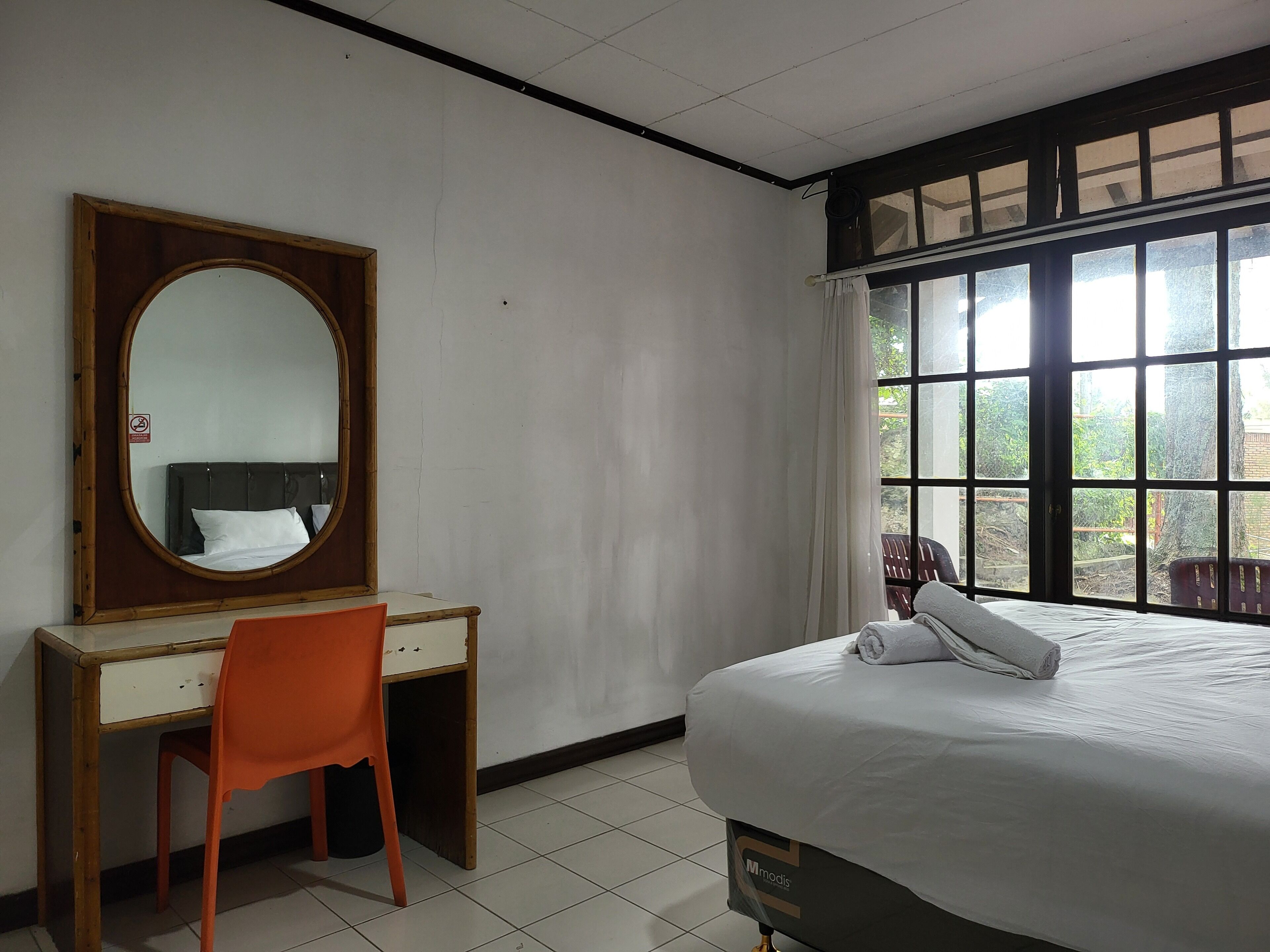 A hotel room with a bed, a desk, a chair, a large mirror, and a window with a view of greenery outside.