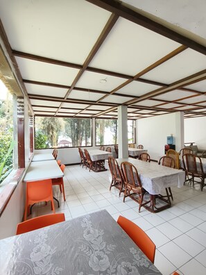 Un espace extérieur abrité, aménagé pour les repas, avec plusieurs tables et chaises, de grandes fenêtres et une vue sur la verdure environnante.
