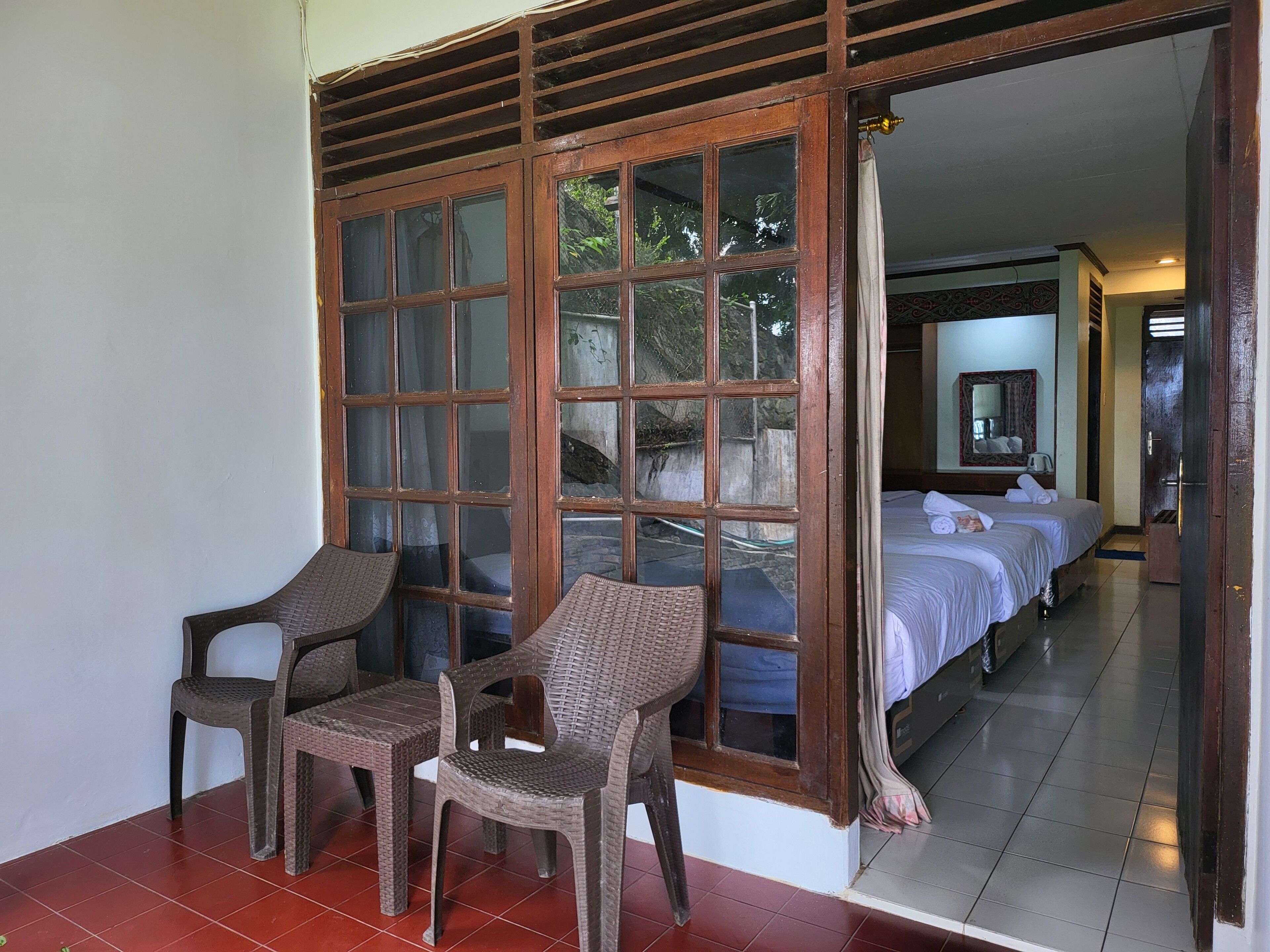 Una habitación con dos sillas de mimbre, una cama con ropa blanca y un ventanal de varios paneles.