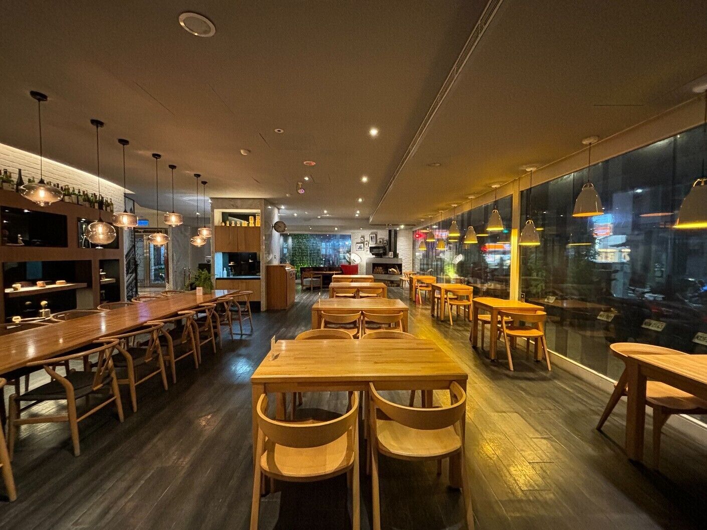 L’intérieur d’un restaurant avec des tables et des chaises en bois, des luminaires suspendus et une vue sur l’extérieur par le biais d’une grande fenêtre.