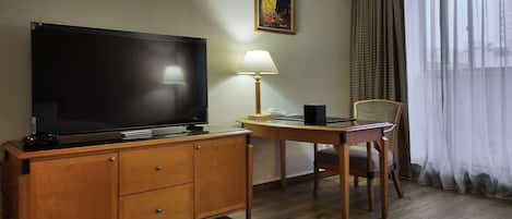 A hotel room with a wooden desk, a television, a lamp, a chair, and a large window with curtains.