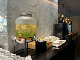Dispensador de agua con un tarro grande lleno de rodajas de fruta y hojas de menta, rodeado de platos y vasos apilados sobre una encimera.