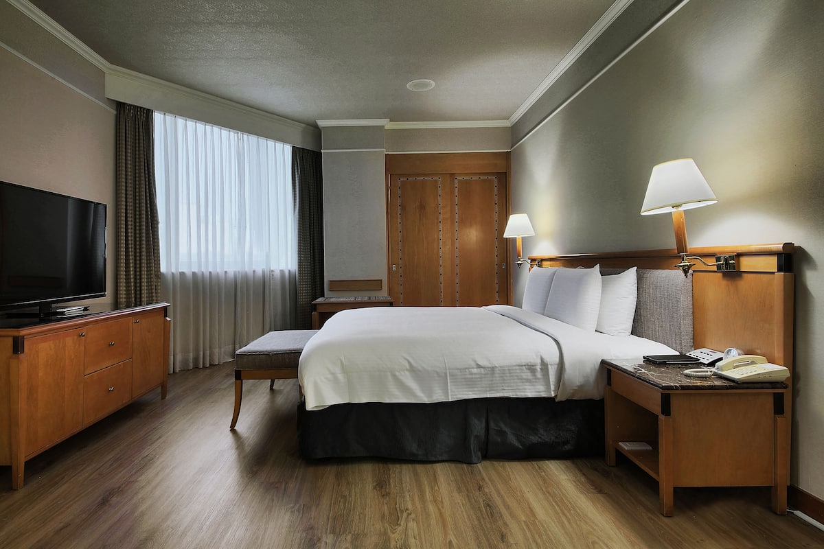 A hotel room with a large bed, bedside lamps, a TV, a wooden dresser, and a window with curtains.