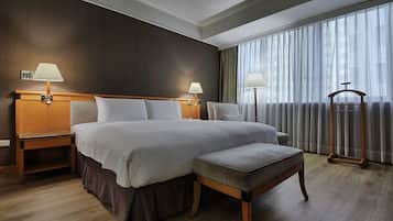 A hotel room with a large bed, two bedside tables with lamps, a wooden bench, and a large window with curtains.