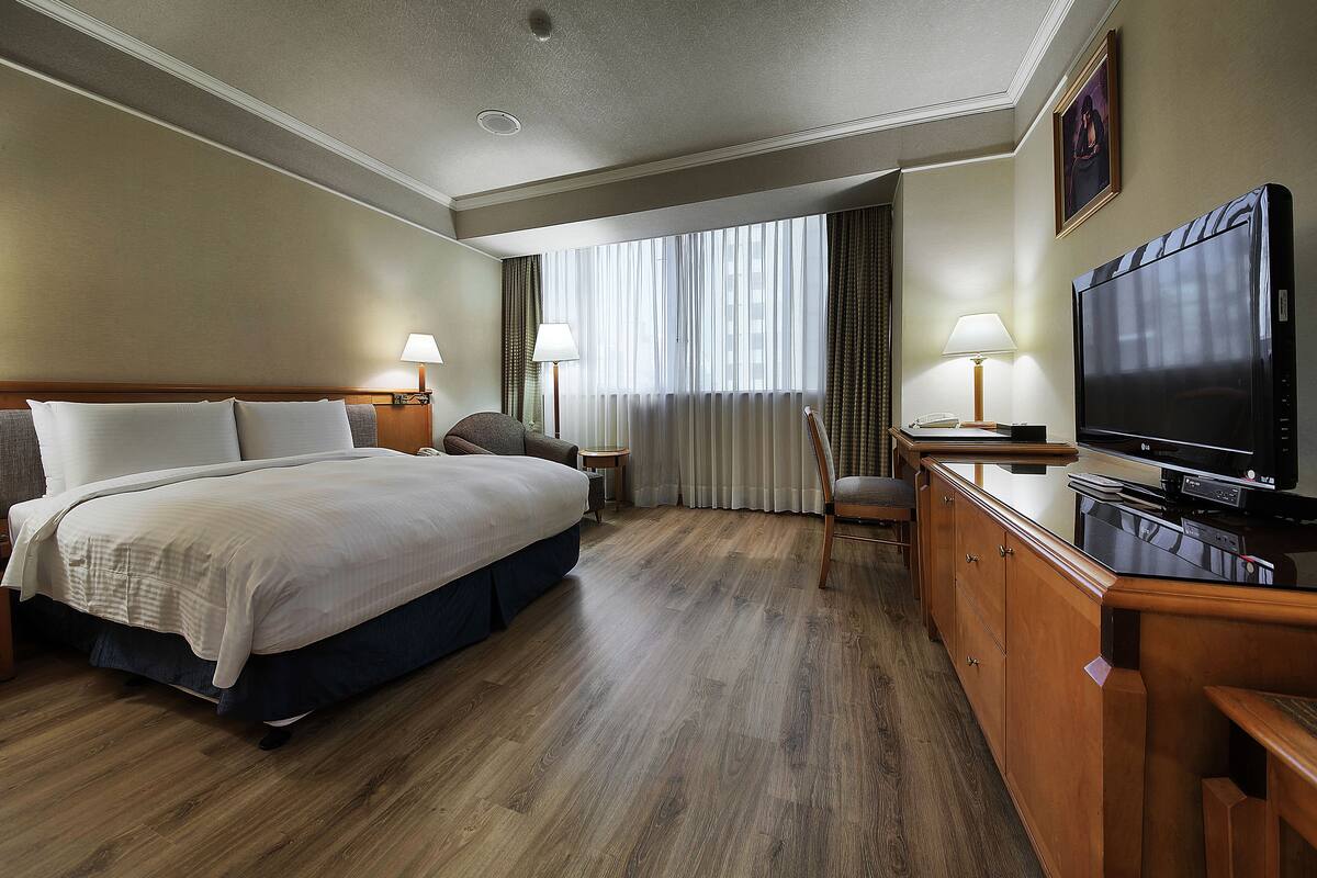 A hotel room with a bed, a desk with a television, a chair, a lamp, and a window with curtains.