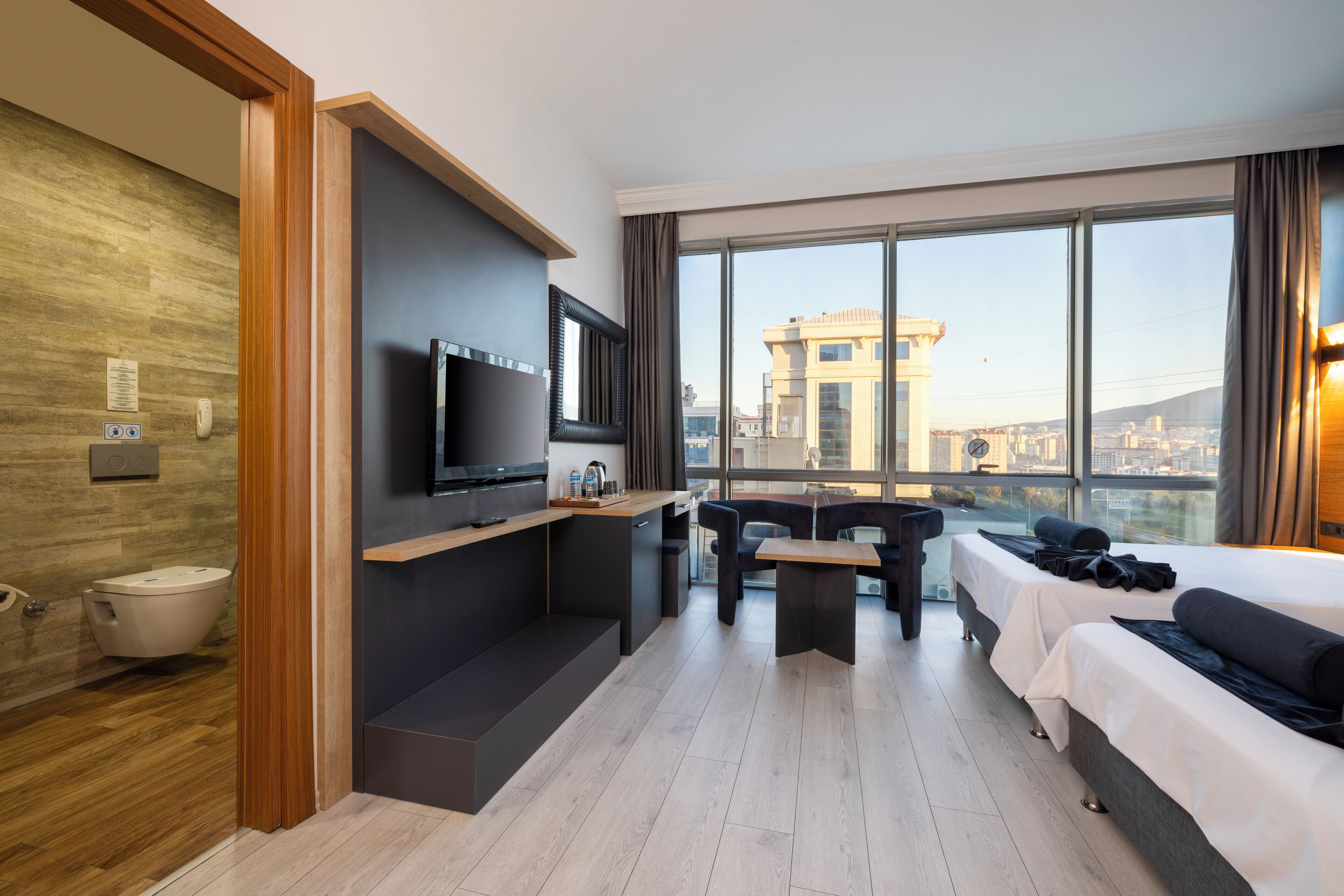 A modern hotel room with a large window, a flat-screen TV, a sofa, a small table, and a kitchenette.