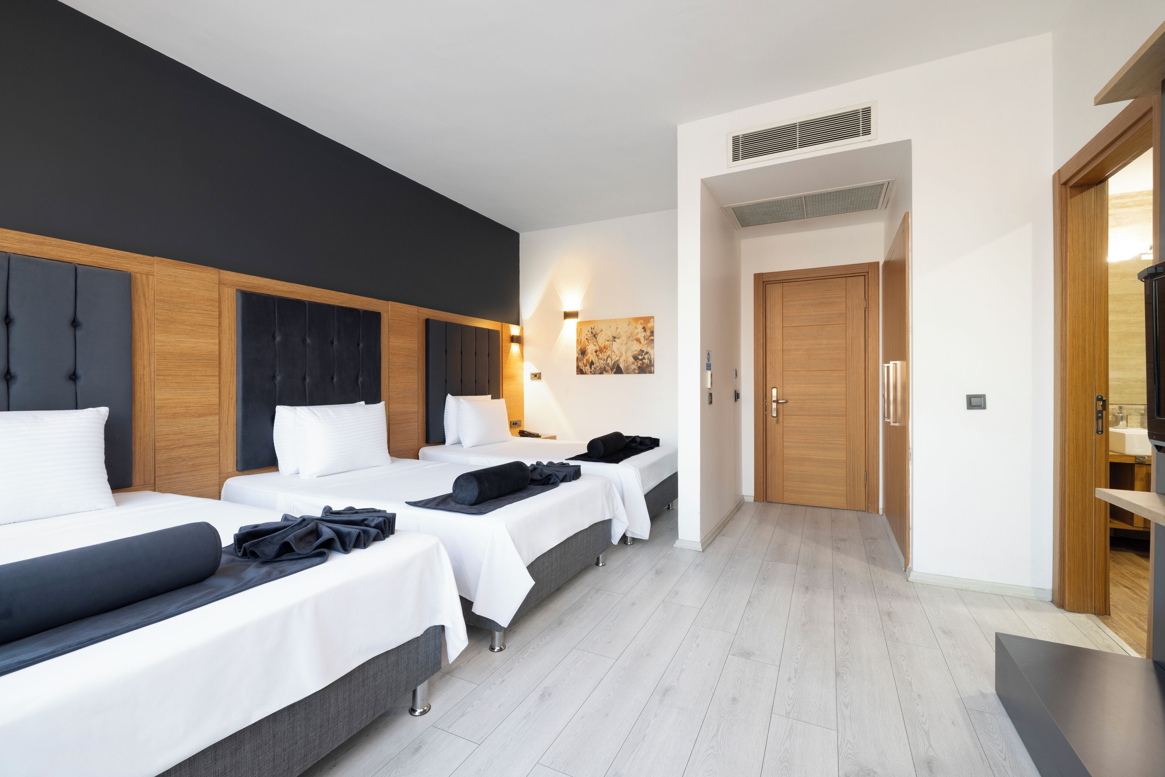 A hotel room with three beds, a wooden wardrobe, a painting on the wall, and a door leading to another room.