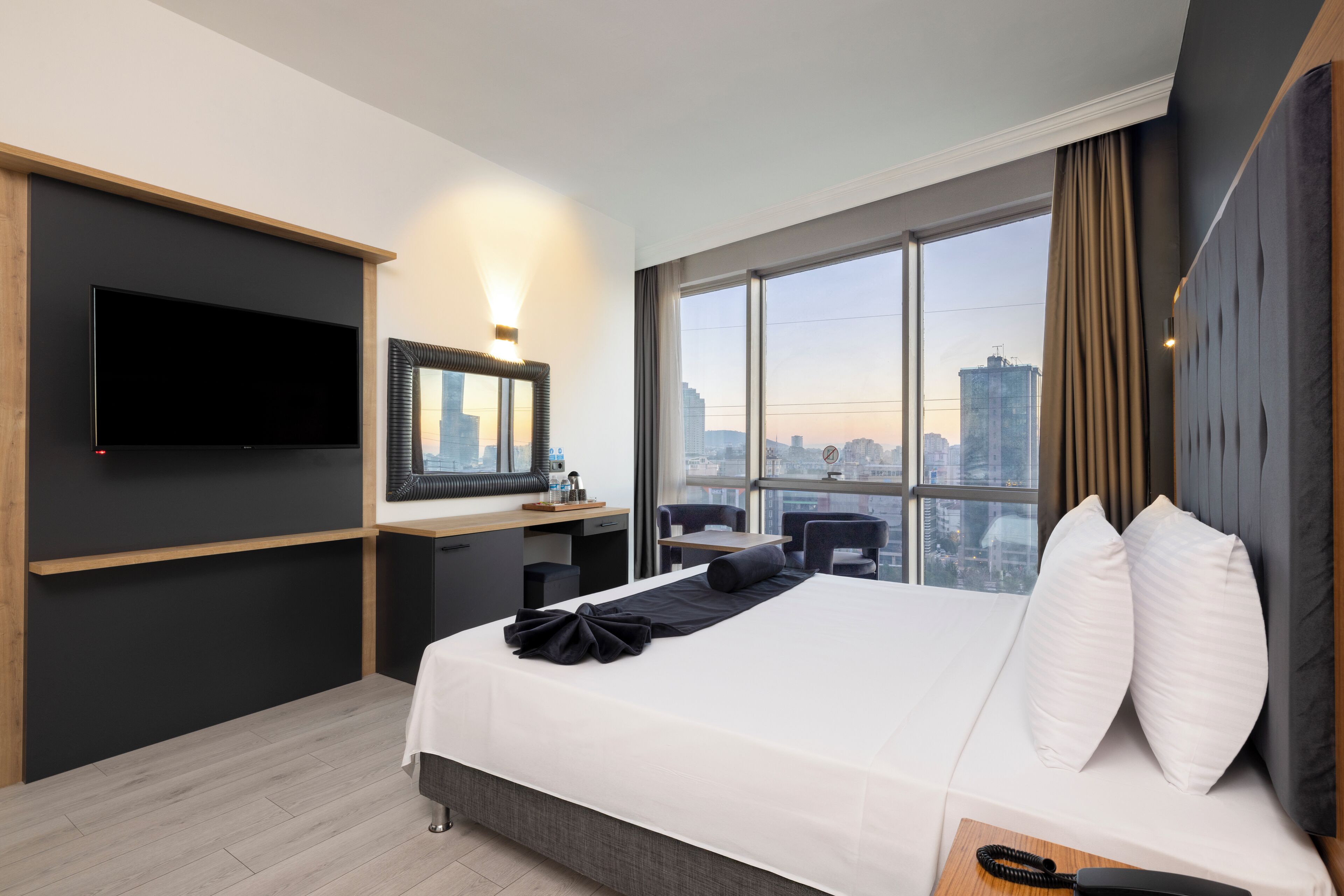 A modern hotel room with a large bed, a desk, a flat-screen TV, and a city view.
