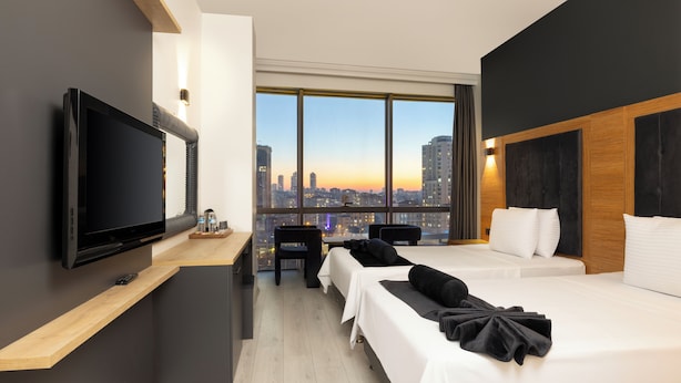 A modern hotel room with a large TV, a desk, two beds, a city view, and a wooden accent wall.