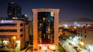 A MyPlus Hotel building with a modern glass facade, surrounded by other buildings and a cityscape in the background.