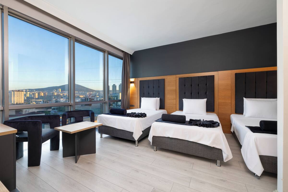 Habitación de hotel con dos camas, un sofá, un escritorio y grandes ventanas con vistas a la ciudad.
