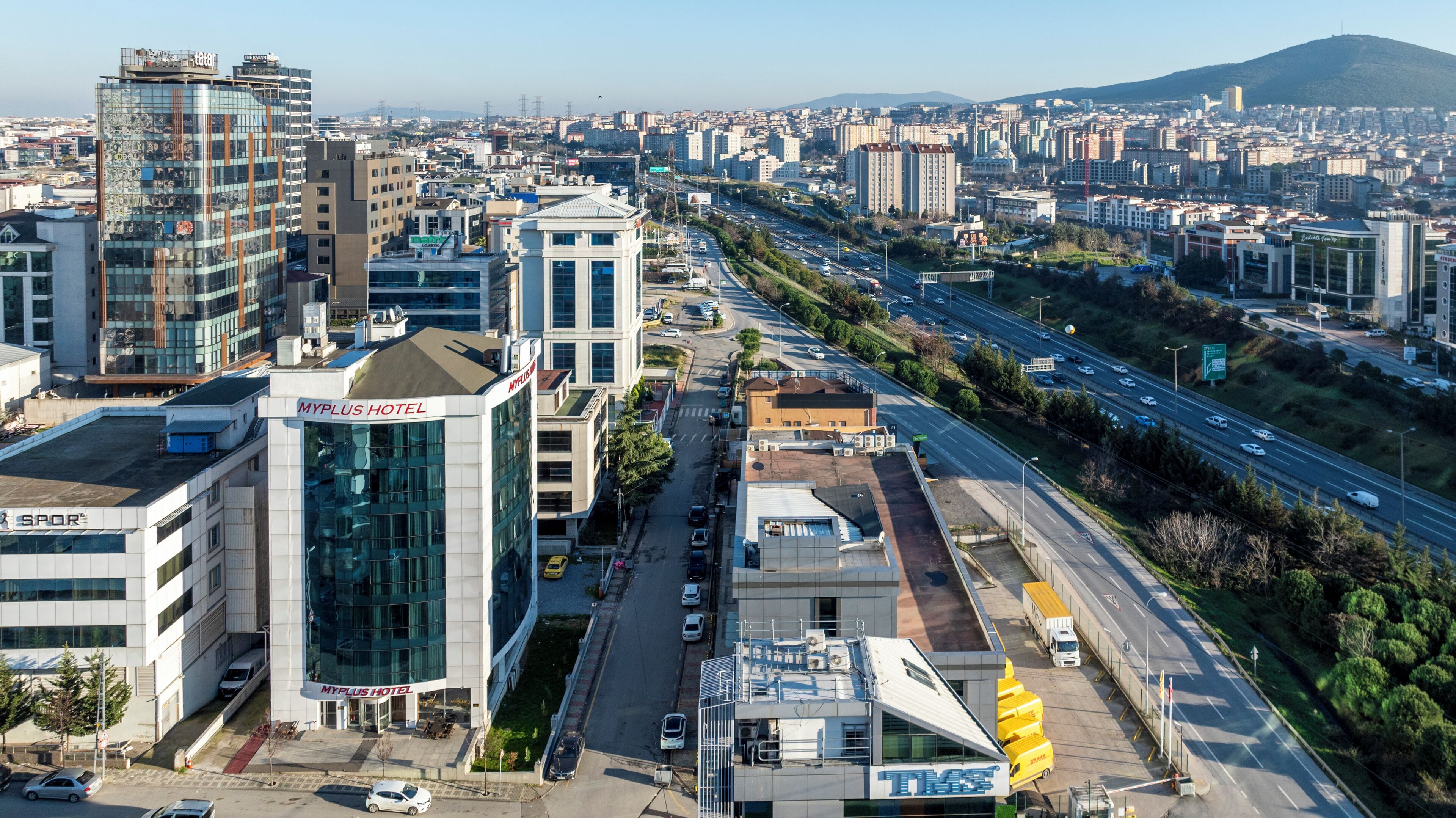 A MyPlus Hotel with a modern glass facade, located near a busy highway and surrounded by urban buildings.