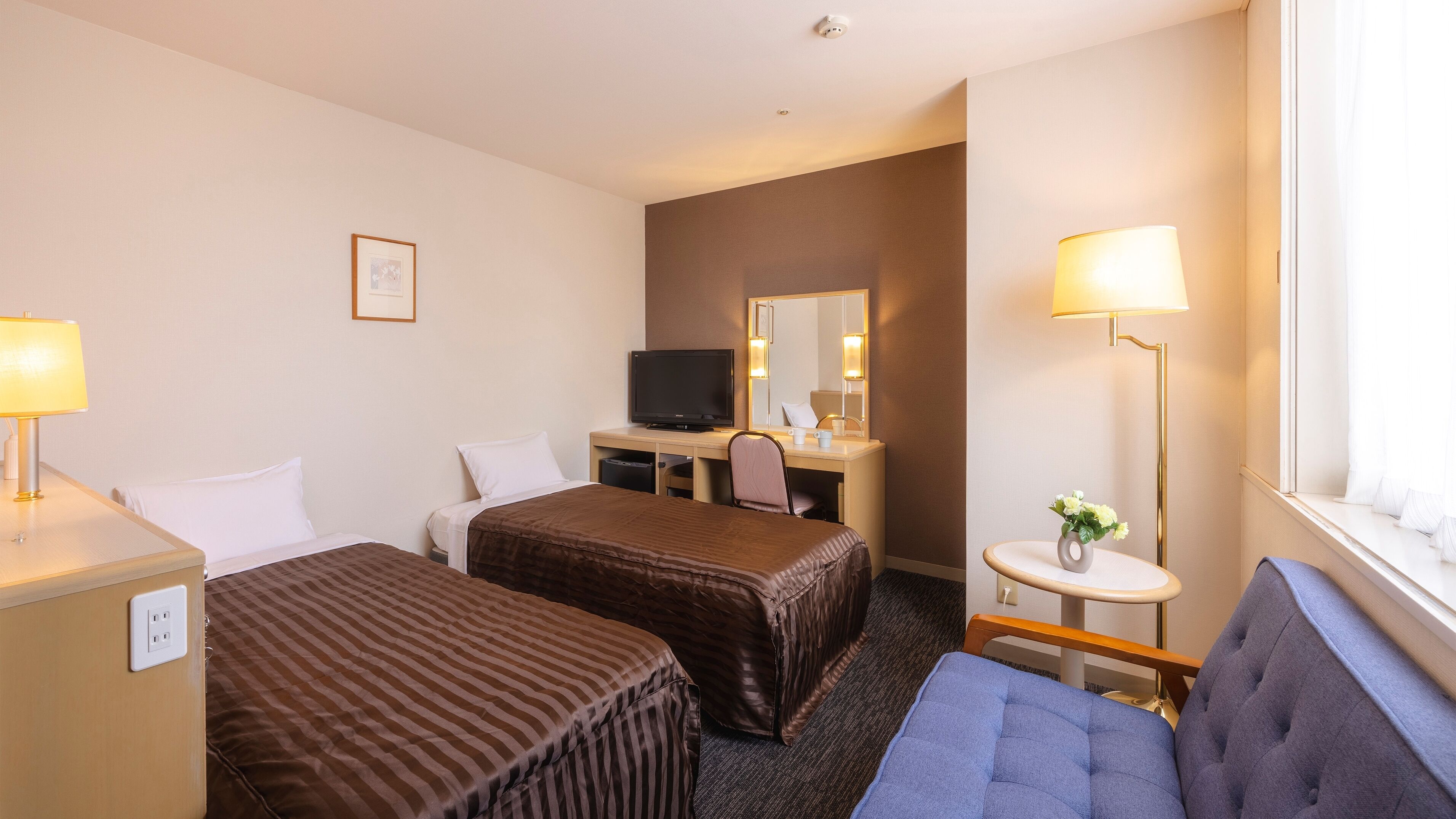 Ein Hotelzimmer mit zwei Betten, einem Schreibtisch mit Fernseher, einem kleinen Tisch mit Blumenvase und einem Fenster mit Vorhängen.
