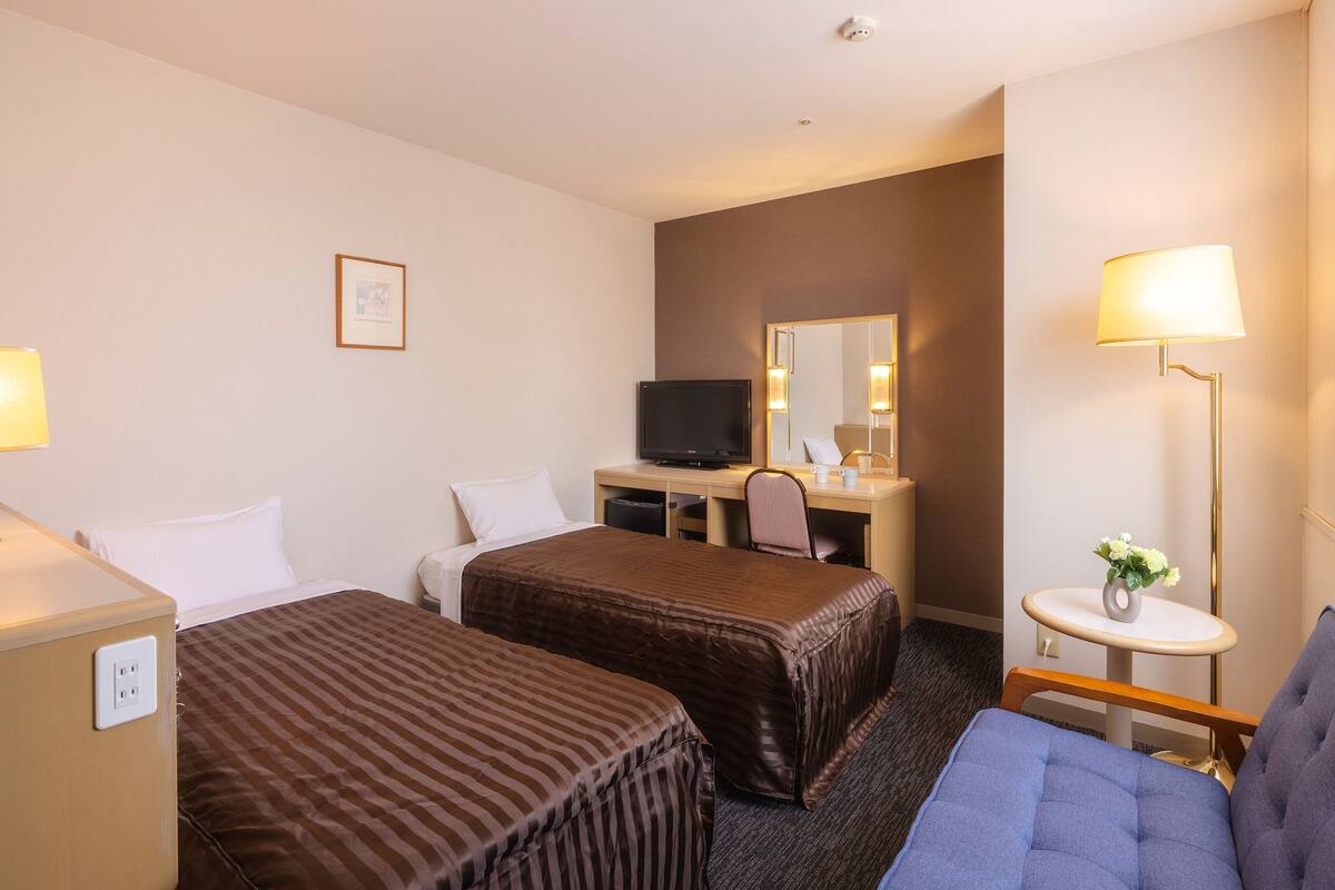 Ein Hotelzimmer mit zwei Betten, einem Schreibtisch mit Fernseher, einem kleinen Tisch mit Blumenvase und einem Fenster mit Vorhängen.