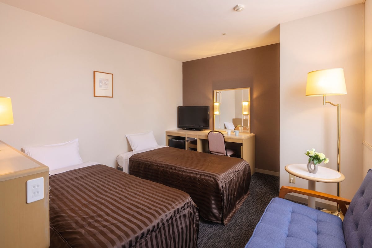 Ein Hotelzimmer mit zwei Betten, einem Schreibtisch mit Fernseher, einem kleinen Tisch mit Blumenvase und einem Fenster mit Vorhängen.