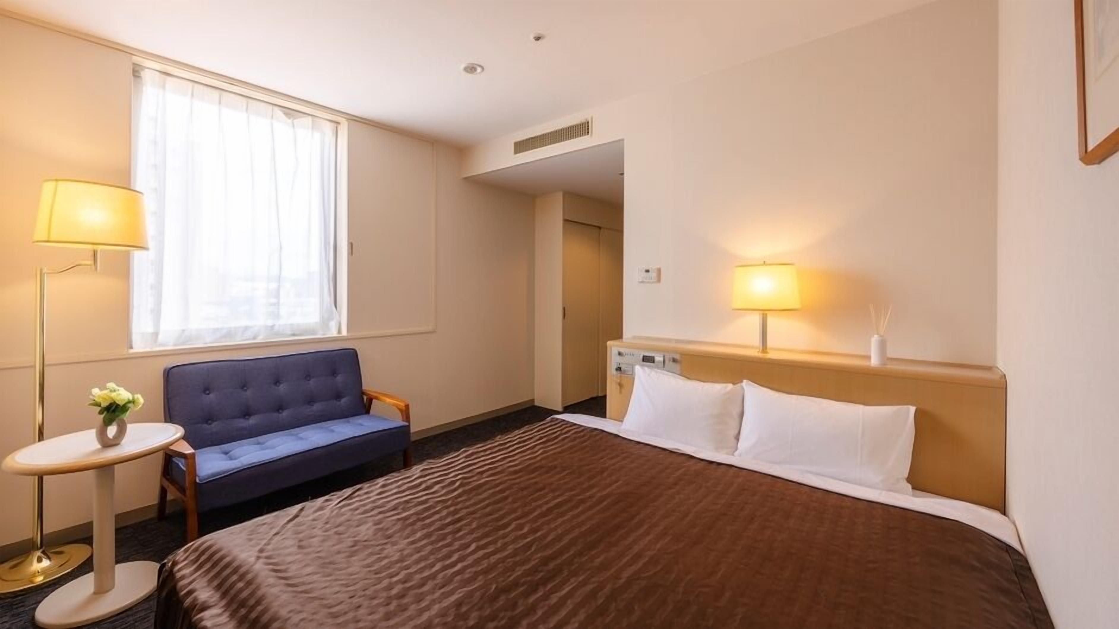 A hotel room with a bed, a sofa, a small table with a lamp, and a window with curtains.
