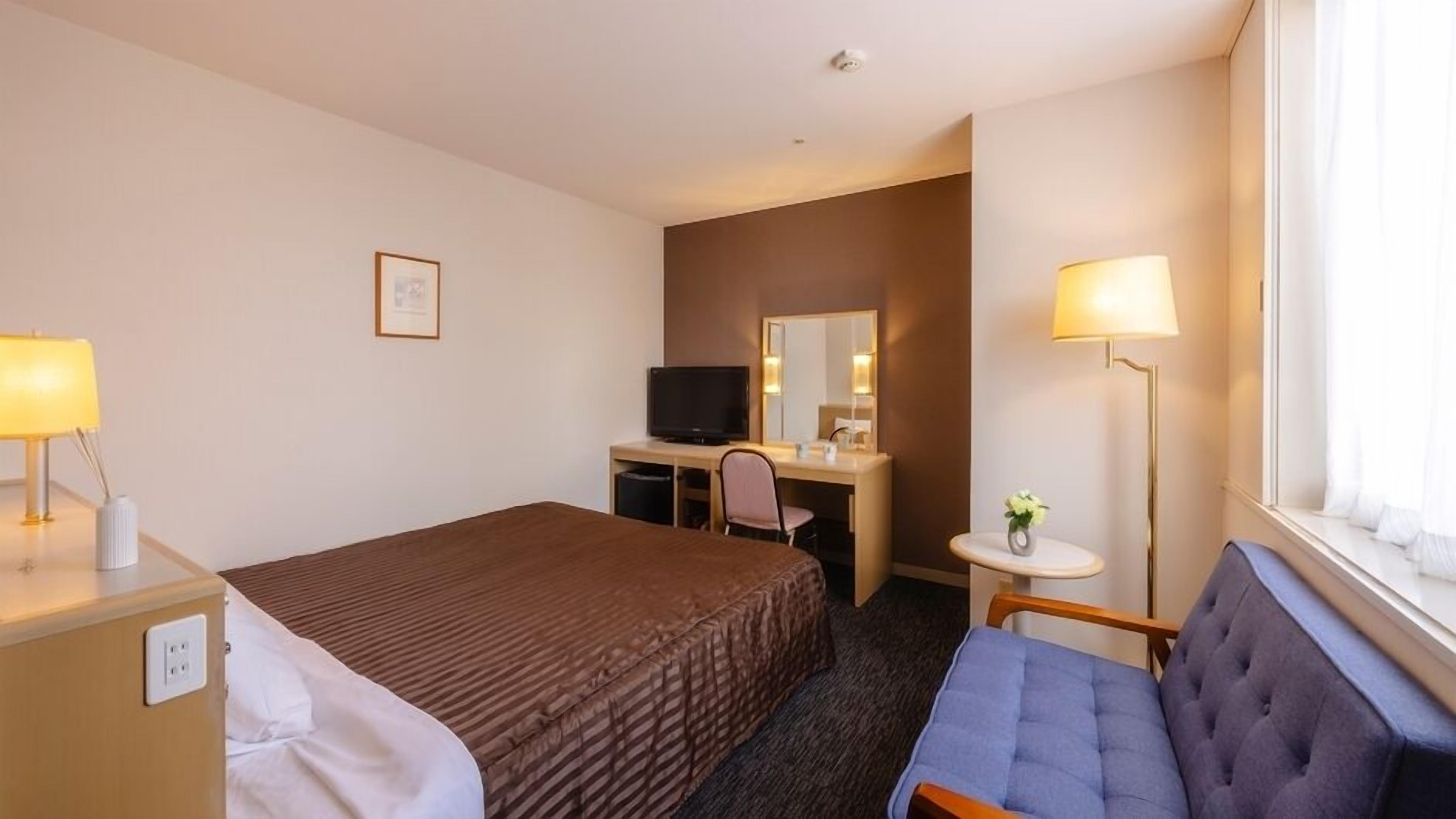A hotel room with a bed, a desk with a television, a chair, a small table with a lamp, and a window with curtains.