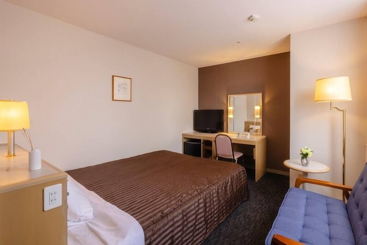 Ein Hotelzimmer mit einem Bett, einem Schreibtisch mit Fernseher, einem Sessel, einem kleinen Tisch mit Lampe und einem Fenster mit Vorhängen.