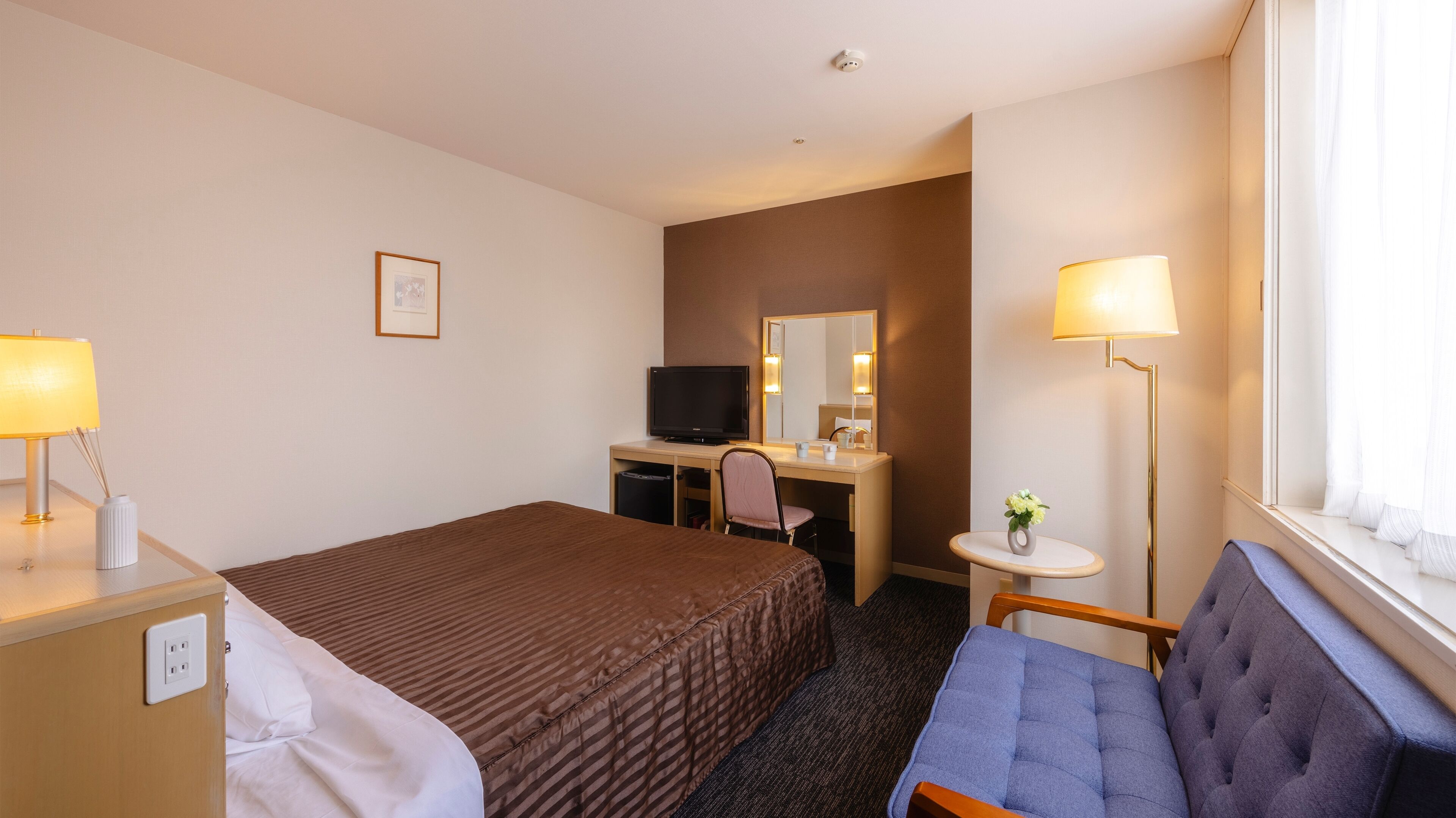Ein Hotelzimmer mit einem Bett, einem Schreibtisch mit Computer, einem Sessel, einem kleinen Tisch mit Lampe und einem Fenster mit Vorhängen.