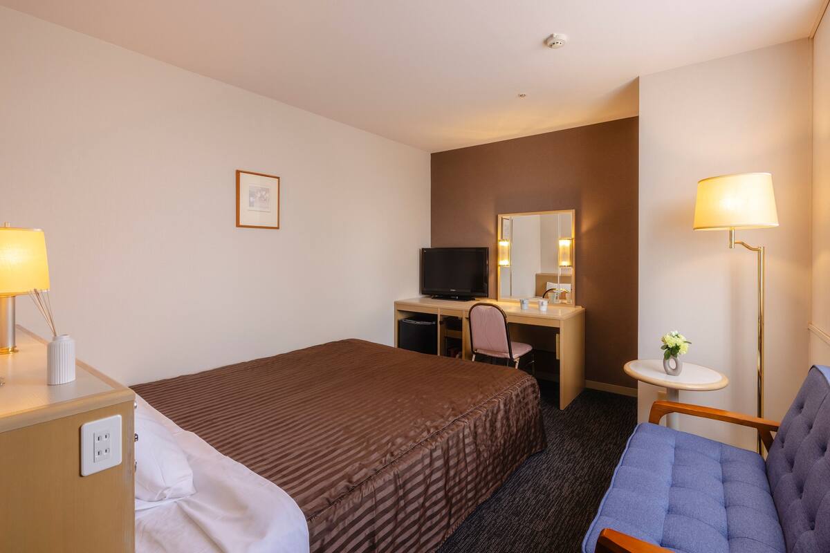 Ein Hotelzimmer mit einem Bett, einem Schreibtisch mit Computer, einem Sessel, einem kleinen Tisch mit Lampe und einem Fenster mit Vorhängen.
