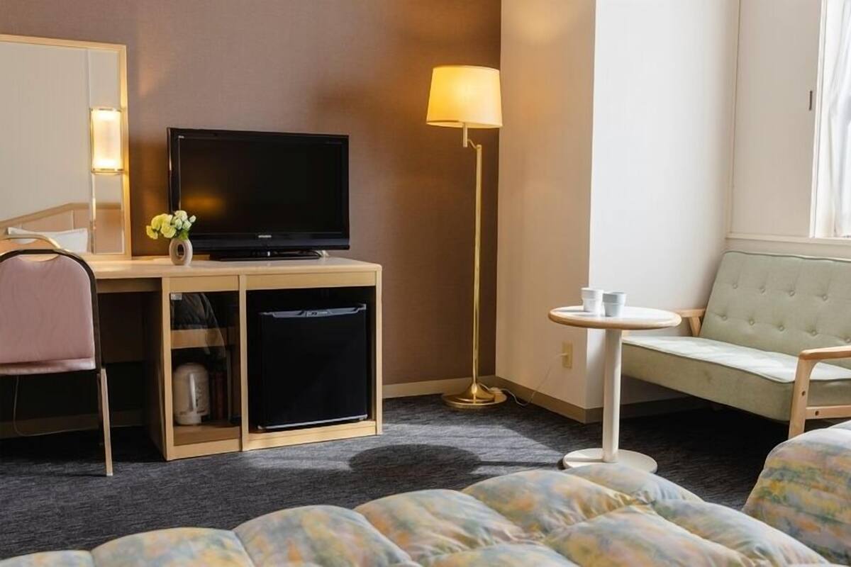 A hotel room with a bed, a desk with a television, a chair, a small table, and a lamp.