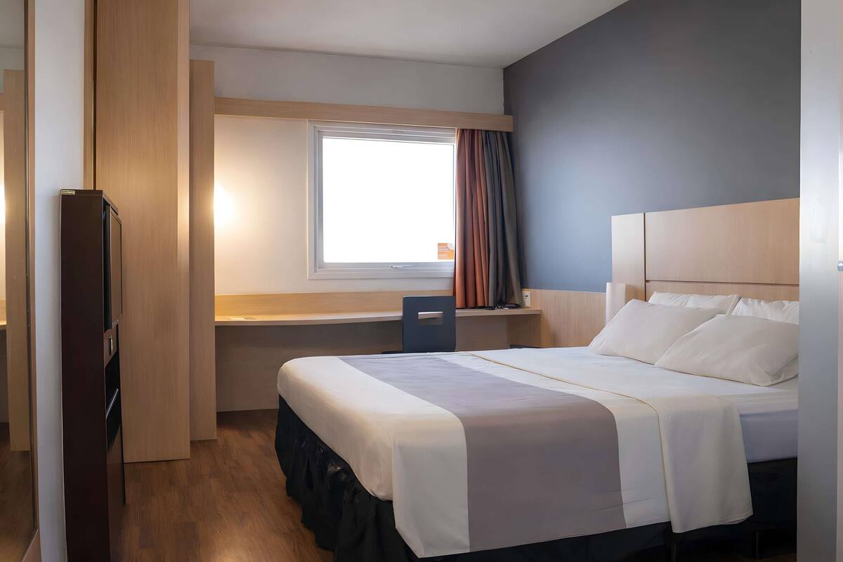 Quarto de hotel com cama, escrivaninha e janela com cortinas.