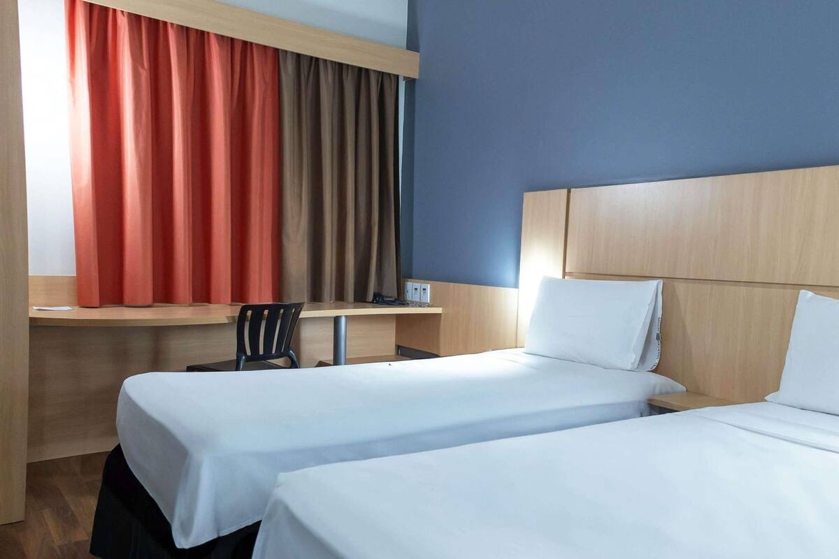 Quarto de hotel com duas camas, uma escrivaninha, uma cadeira e uma janela com cortinas.