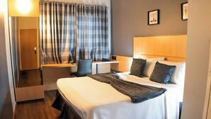 Una habitación de hotel con cama, mesita de noche, lámpara y ventana con cortinas.