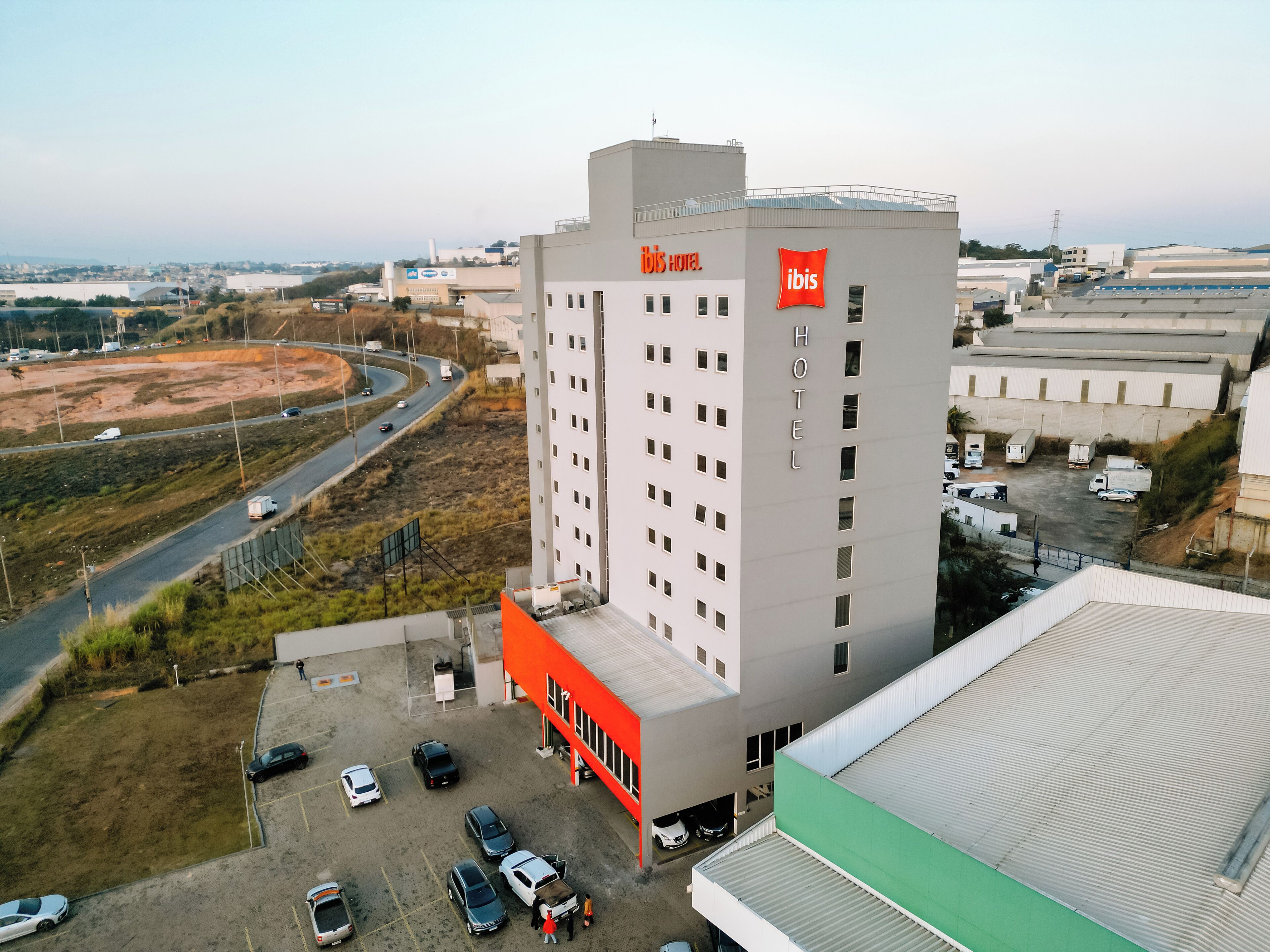 Un hotel moderno con un cartello ben visibile con il marchio "ibis", diversi posti auto e vista sull'autostrada e il paesaggio circostante.