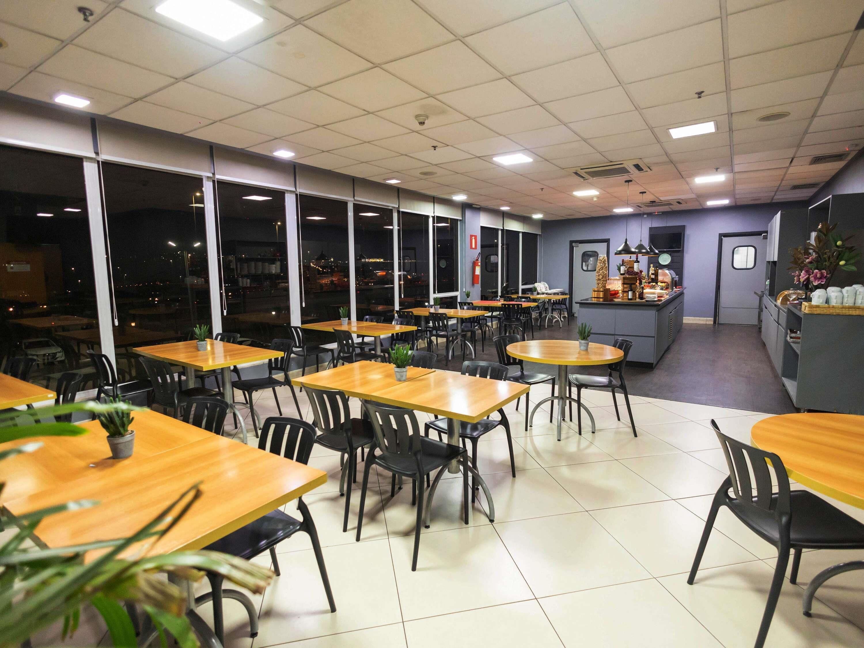 Um restaurante com várias mesas e cadeiras, um balcão com vitrine e grandes janelas.