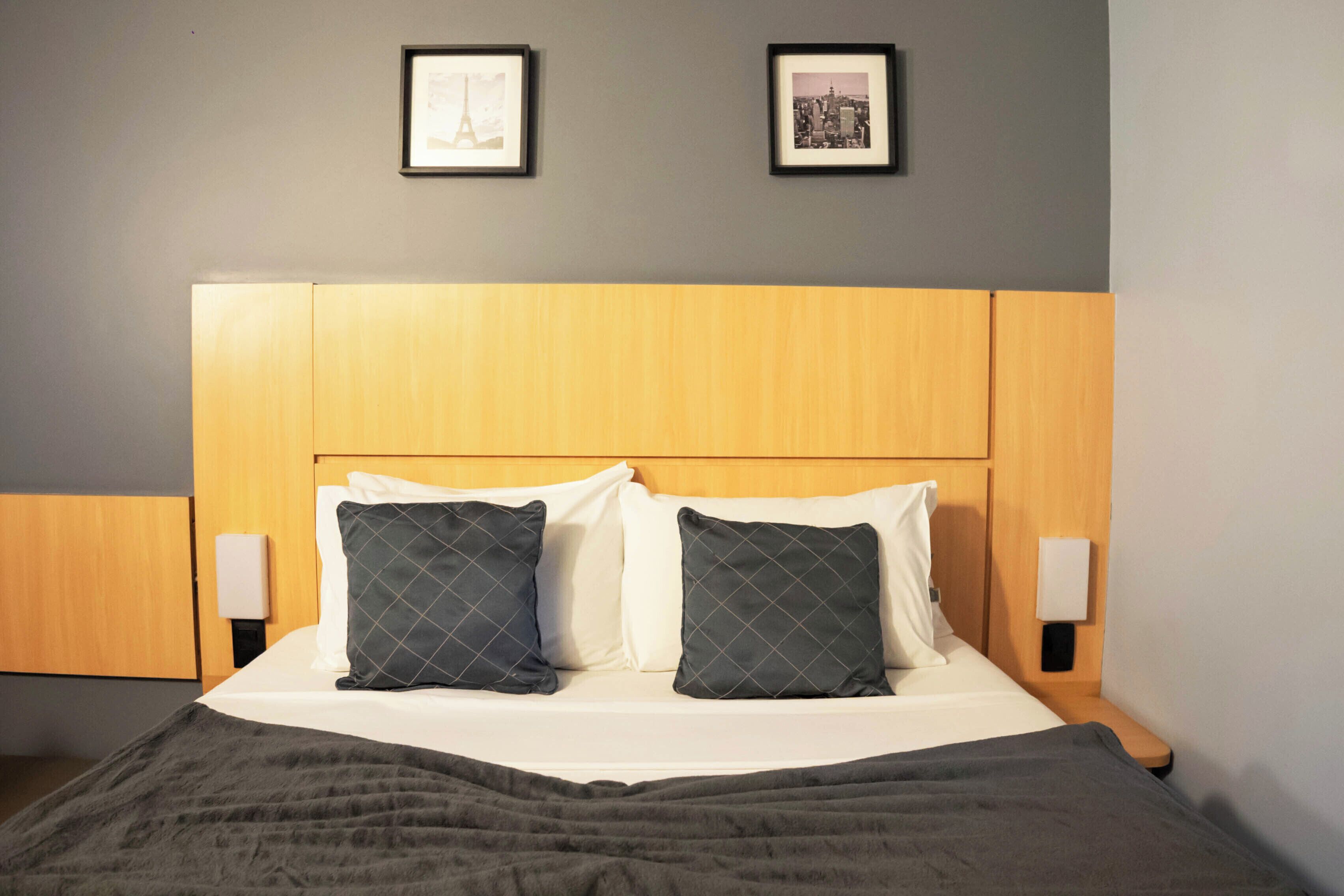 Cama bem arrumada com cabeceira de madeira, duas mesas de cabeceira com abajures e duas quadros na parede.
