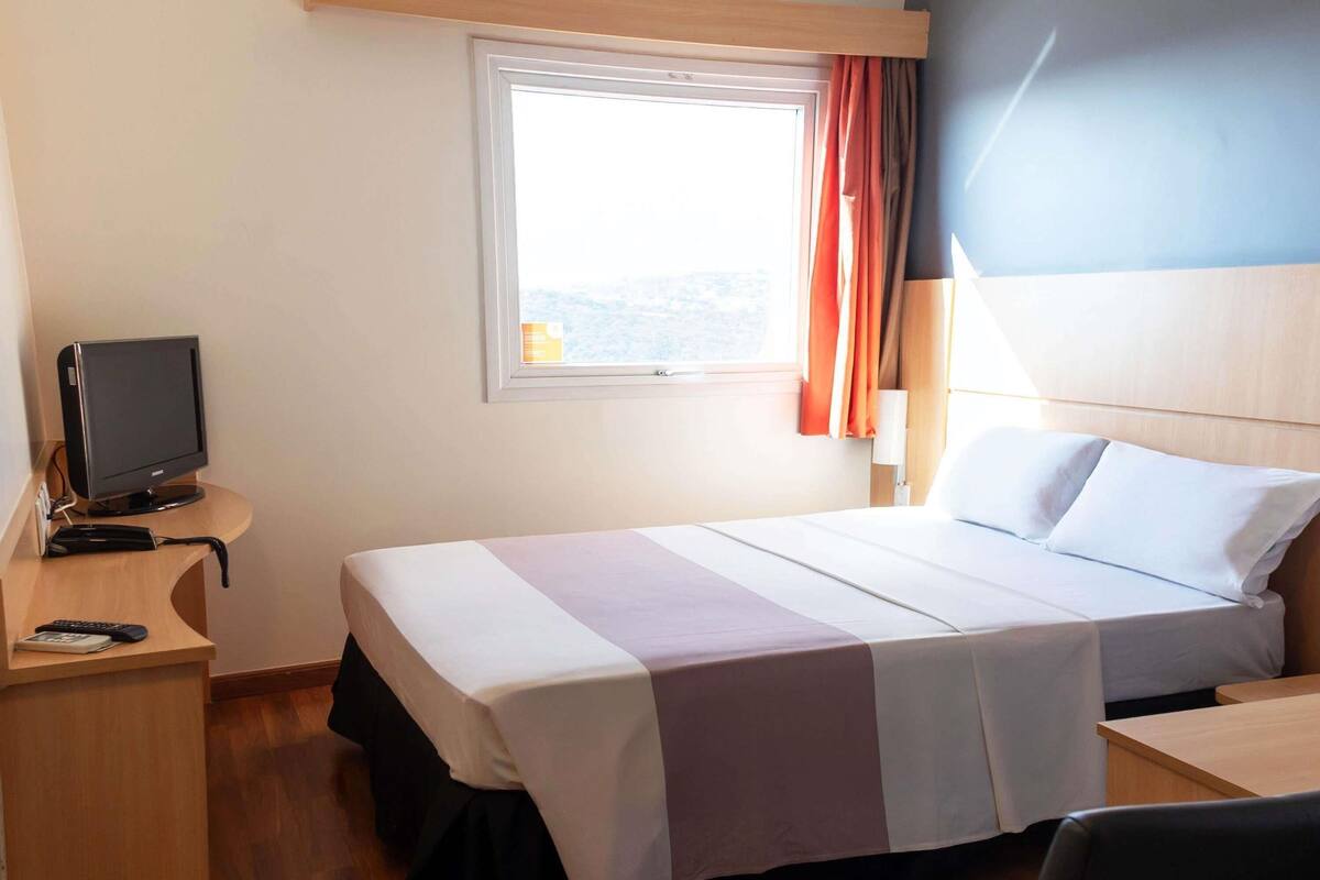 Quarto de hotel com cama, escrivaninha, televisão e janela com cortinas.