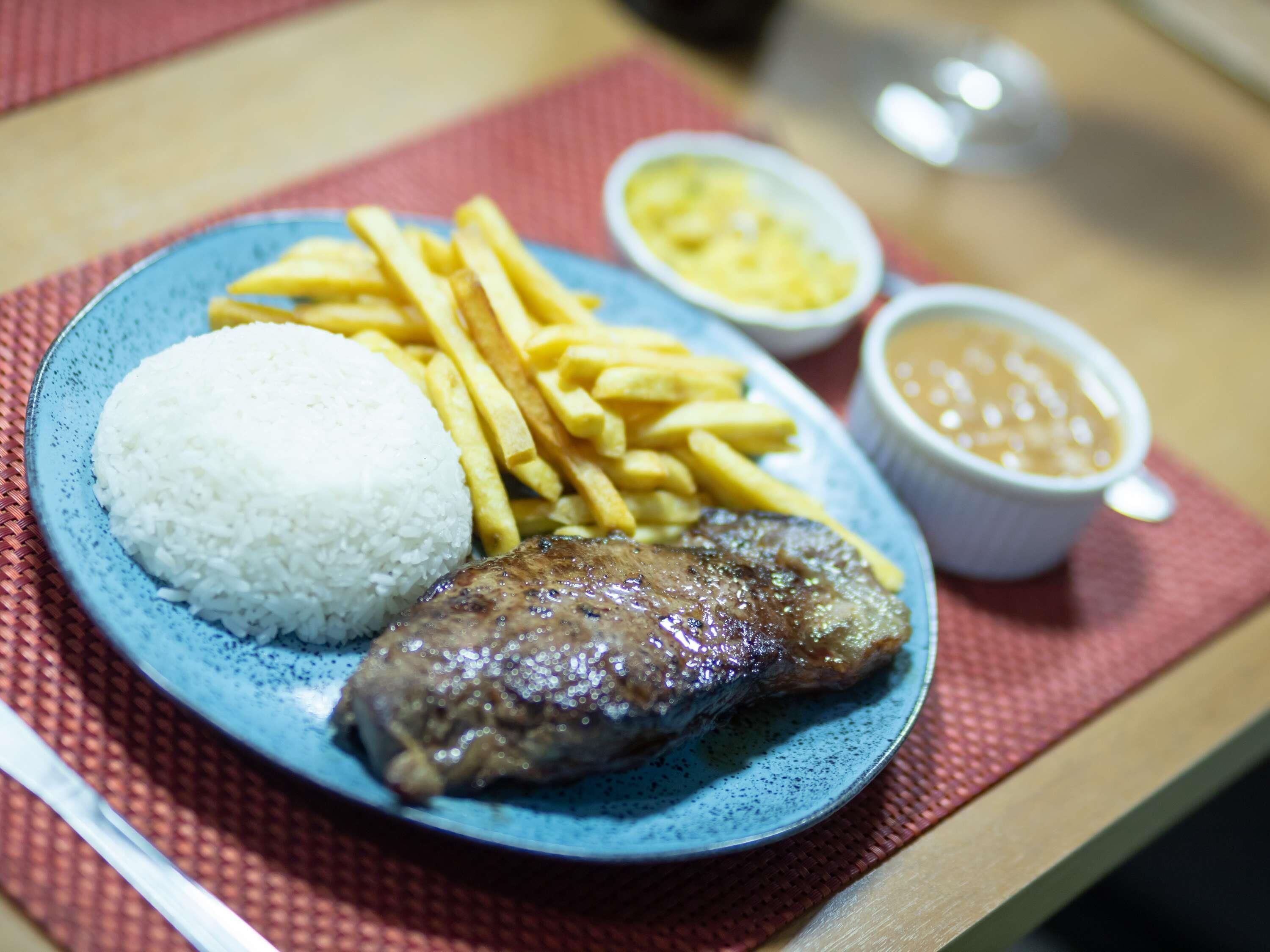 A plate with rice, fries, a grilled meat, and two small bowls of sauce on a table.