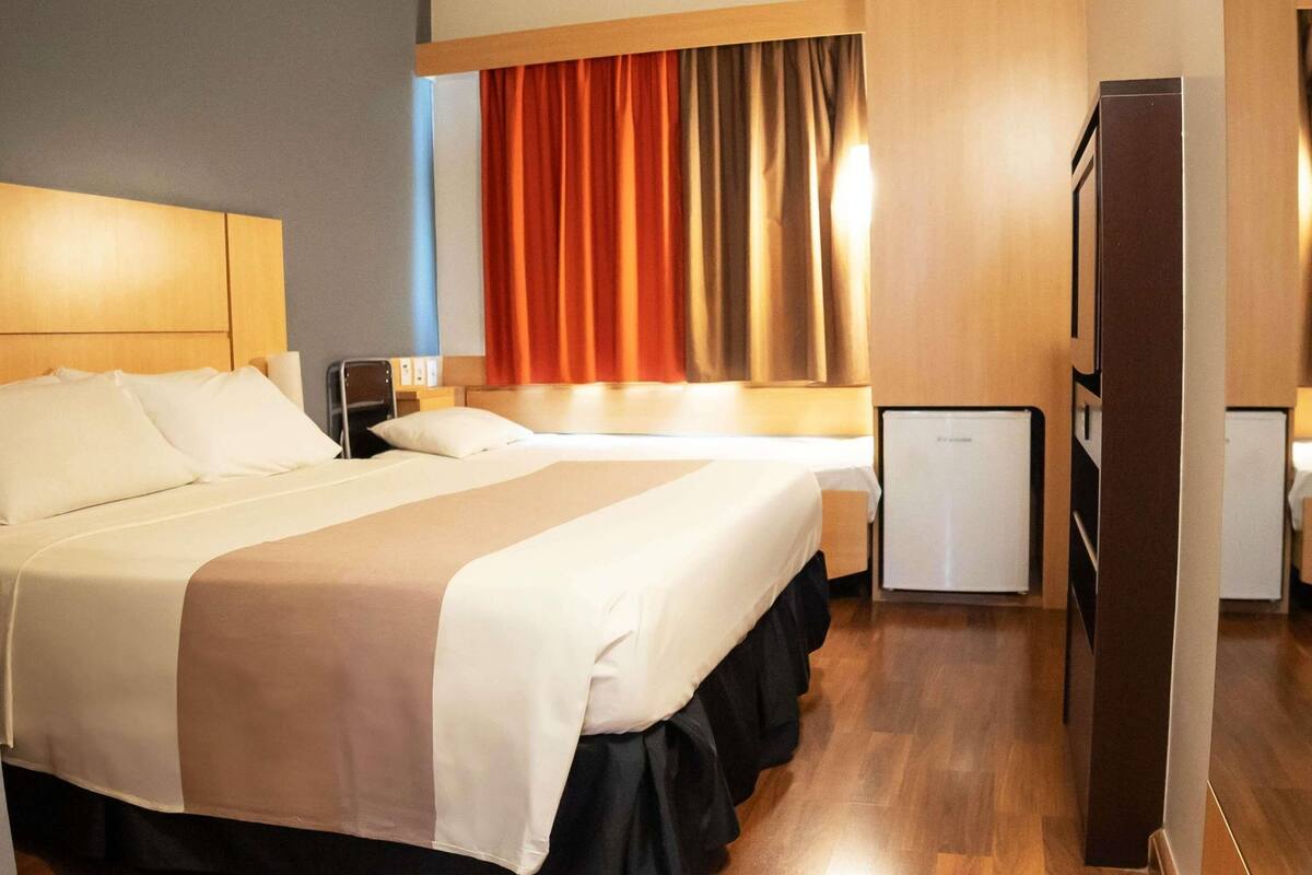 Quarto de hotel com duas camas, um pequeno frigorífico e um guarda-roupa de madeira.