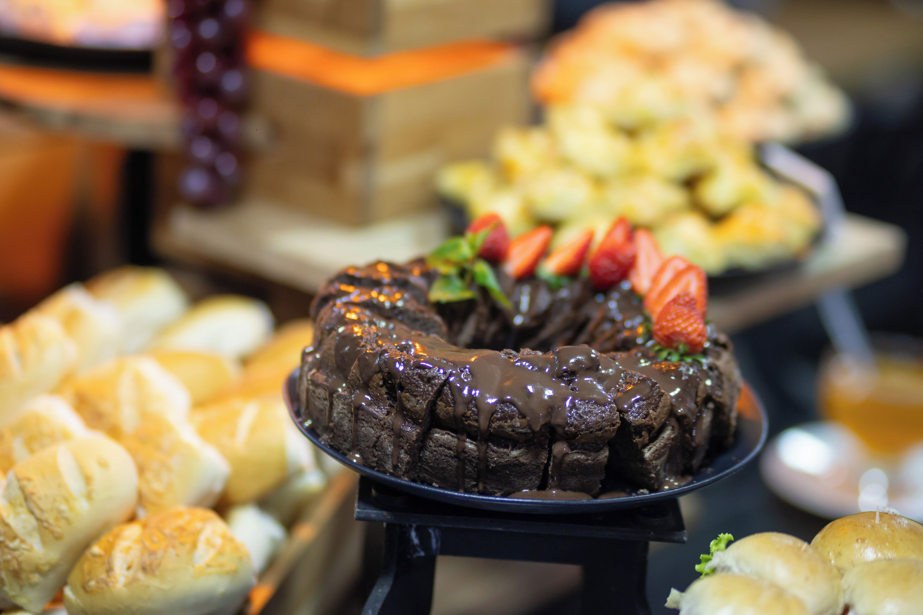 A dessert table with a chocolate cake topped with strawberries, surrounded by various pastries and bread.