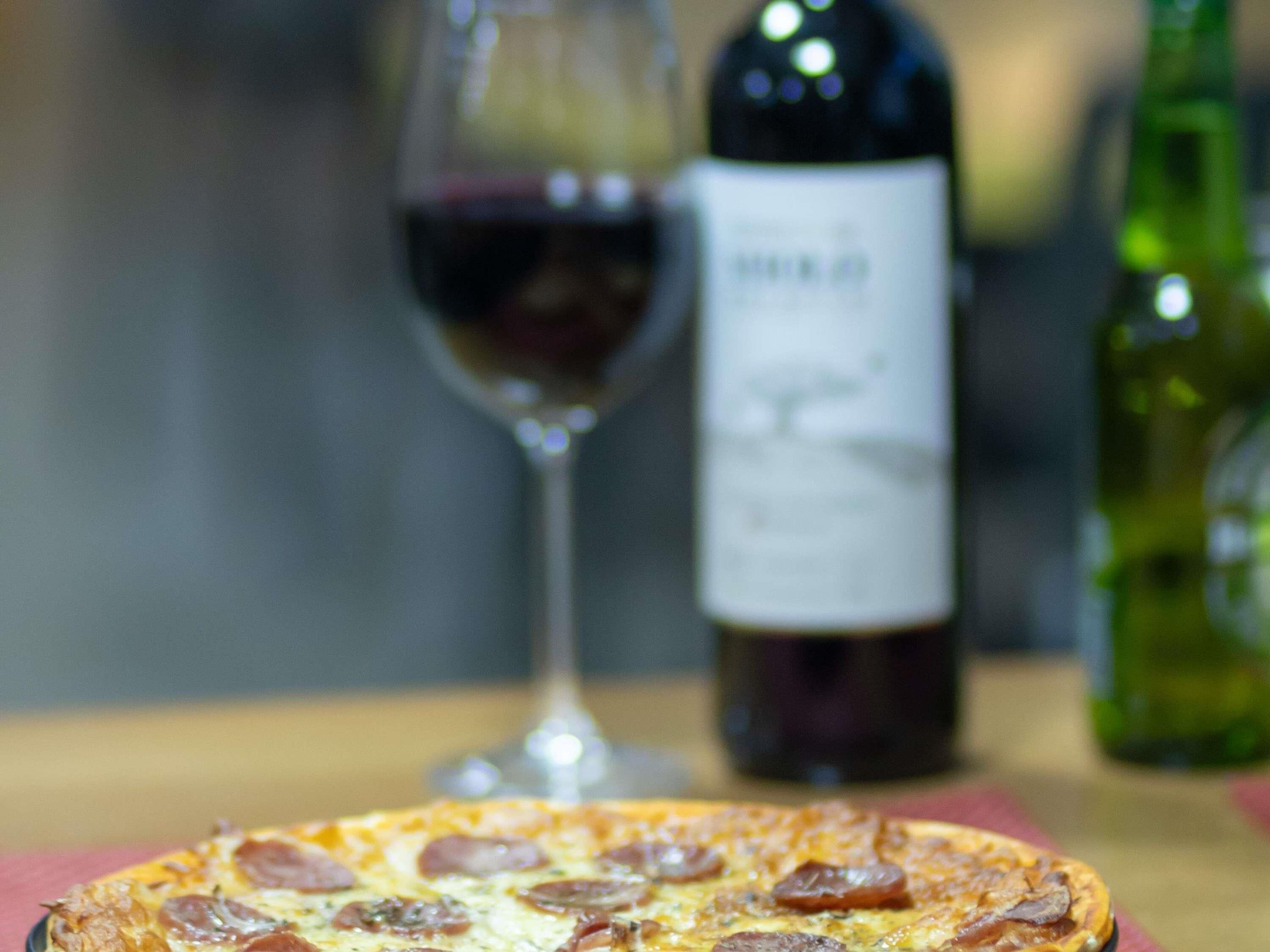 Una pizza sobre la mesa, con una botella de vino y una copa al fondo.