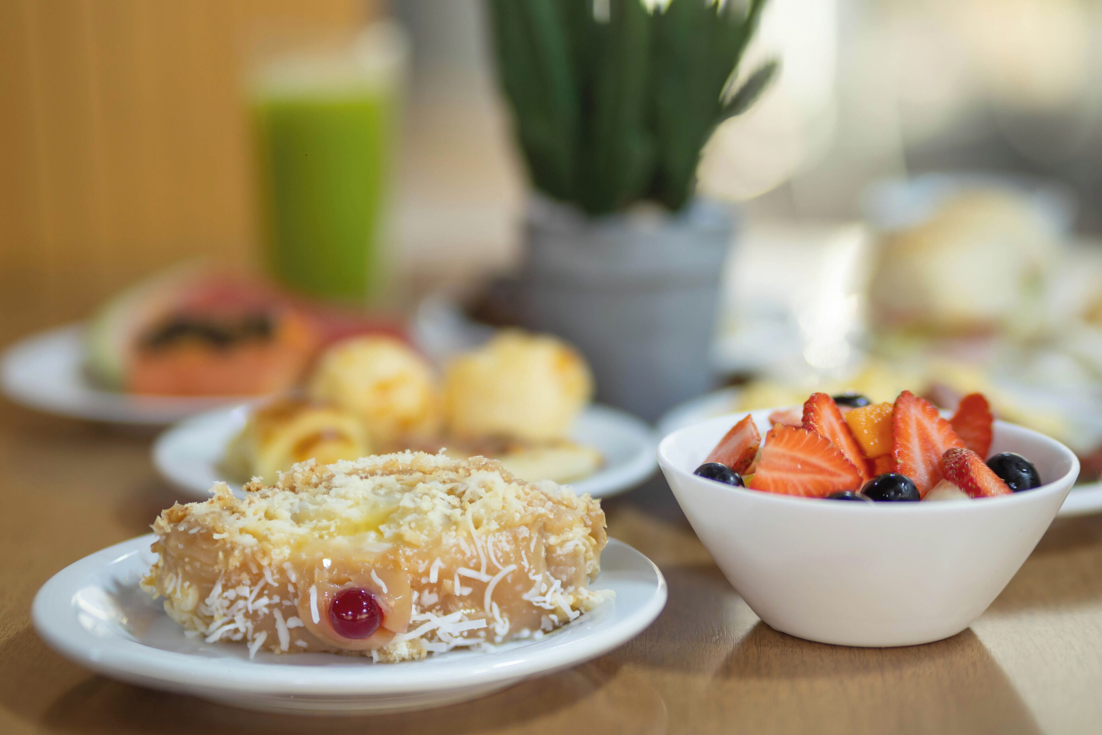 Uma mesa com diversos alimentos, incluindo um pastel com coco ralado, uma tigela de frutas vermelhas e outros pratos ao fundo.