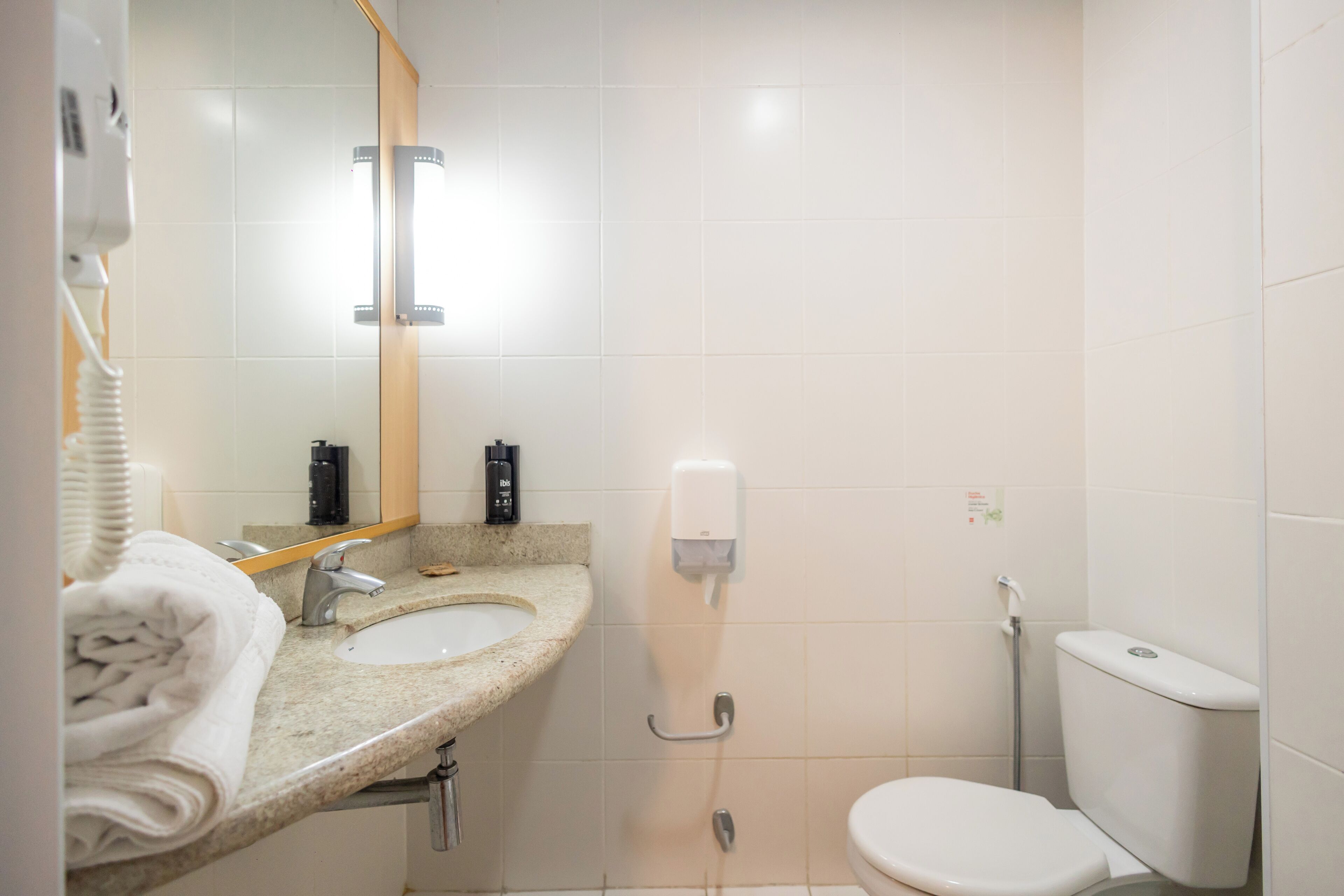 A bathroom with a sink, mirror, towel, toilet, and soap dispenser.