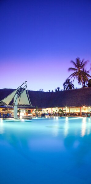 A pool area with a thatched-roof structure, palm trees, and a well-lit lounge area. - Turtle Bay Beach Club (Watamu)