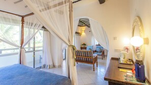 A room with a bed, a chair, a table, and a mirror. There is a window with curtains and a view of palm trees outside. - Turtle Bay Beach Club (Watamu)