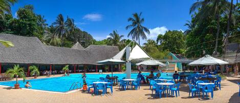 A pool area with blue chairs and tables, surrounded by palm trees and thatched-roof buildings.