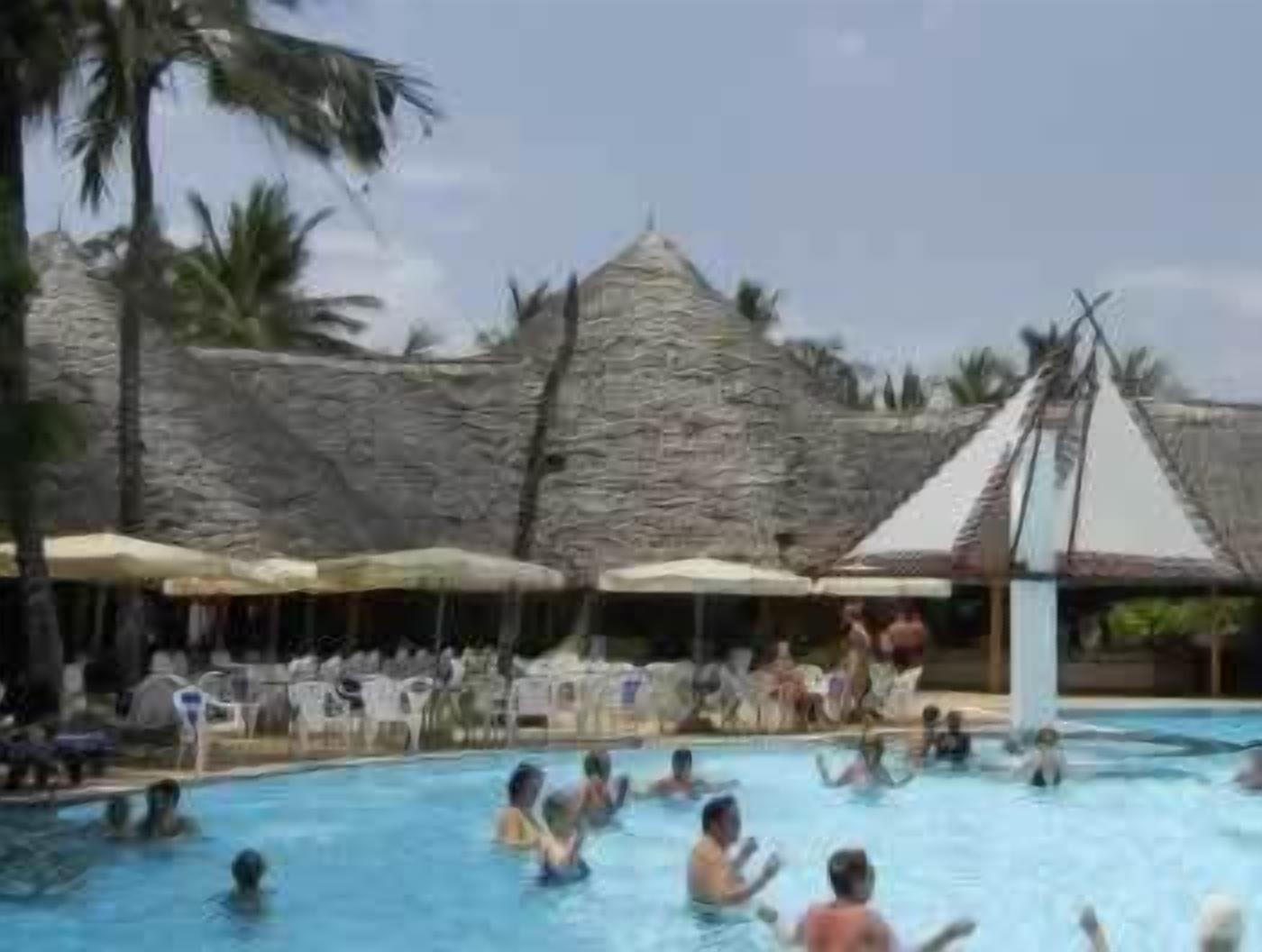 A swimming pool area with people swimming and sunbathing, surrounded by palm trees and thatched-roof structures.