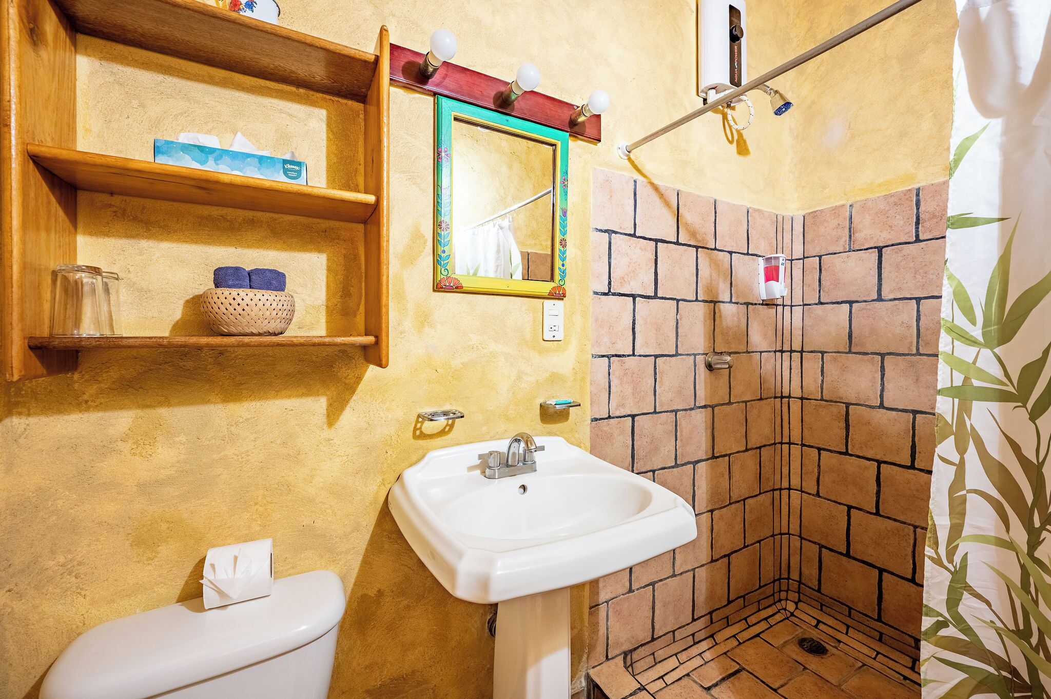 A bathroom with a white sink, a toilet, a shower with a brick wall, and a shelf with toiletries.