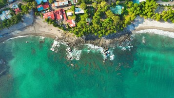 An aerial view of a coastal area with buildings, a beach, and clear turquoise waters.