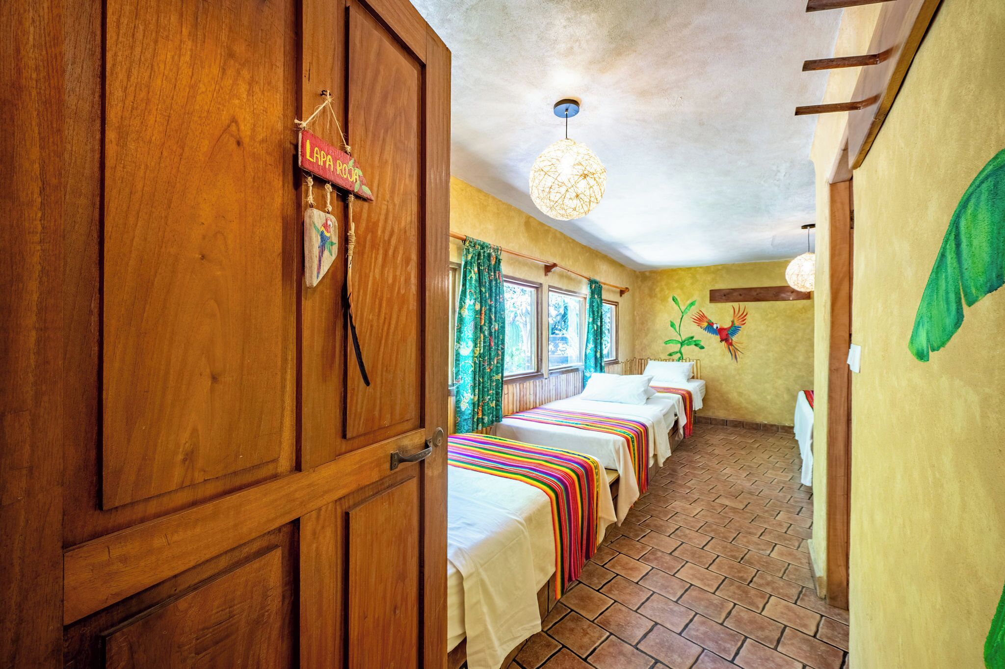Quarto com paredes de madeira, chão de azulejos, cama com manta listrada e colorida, luminária pendente e um adorno na parede.