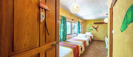 Quarto com paredes de madeira, chão de azulejos, cama com manta listrada e colorida, luminária pendente e um adorno na parede.